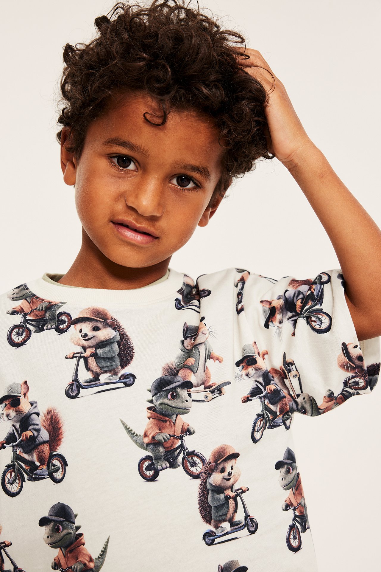 Närbild framifrån: Barn i offwhite bomulls-t-shirt med heltäckande djurmönster av cyklande djur och skateboards.
