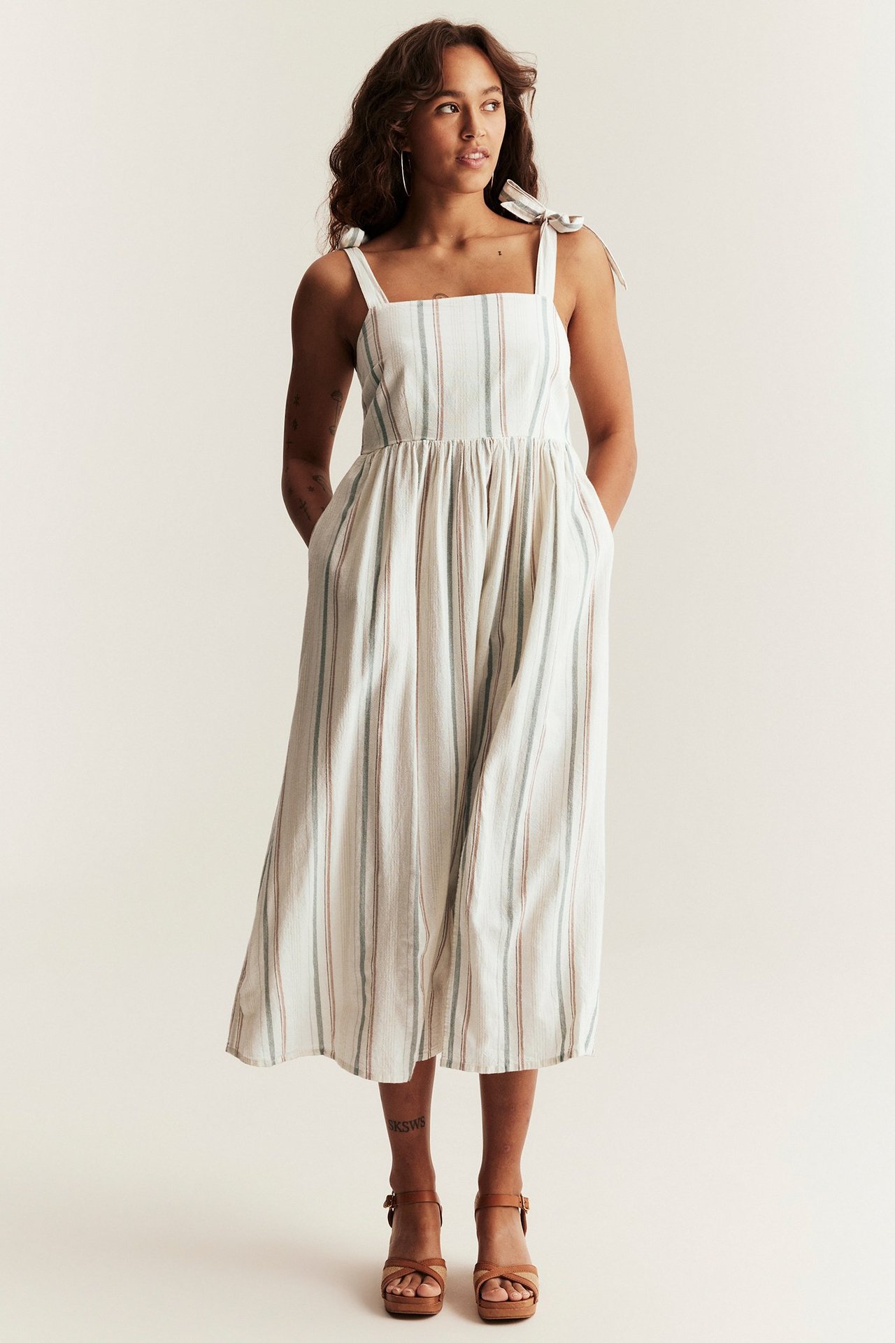 Framifrån: kvinna i randig klänning i offwhite bomull med knytband, smockrygg, och bruna platåsandaler.