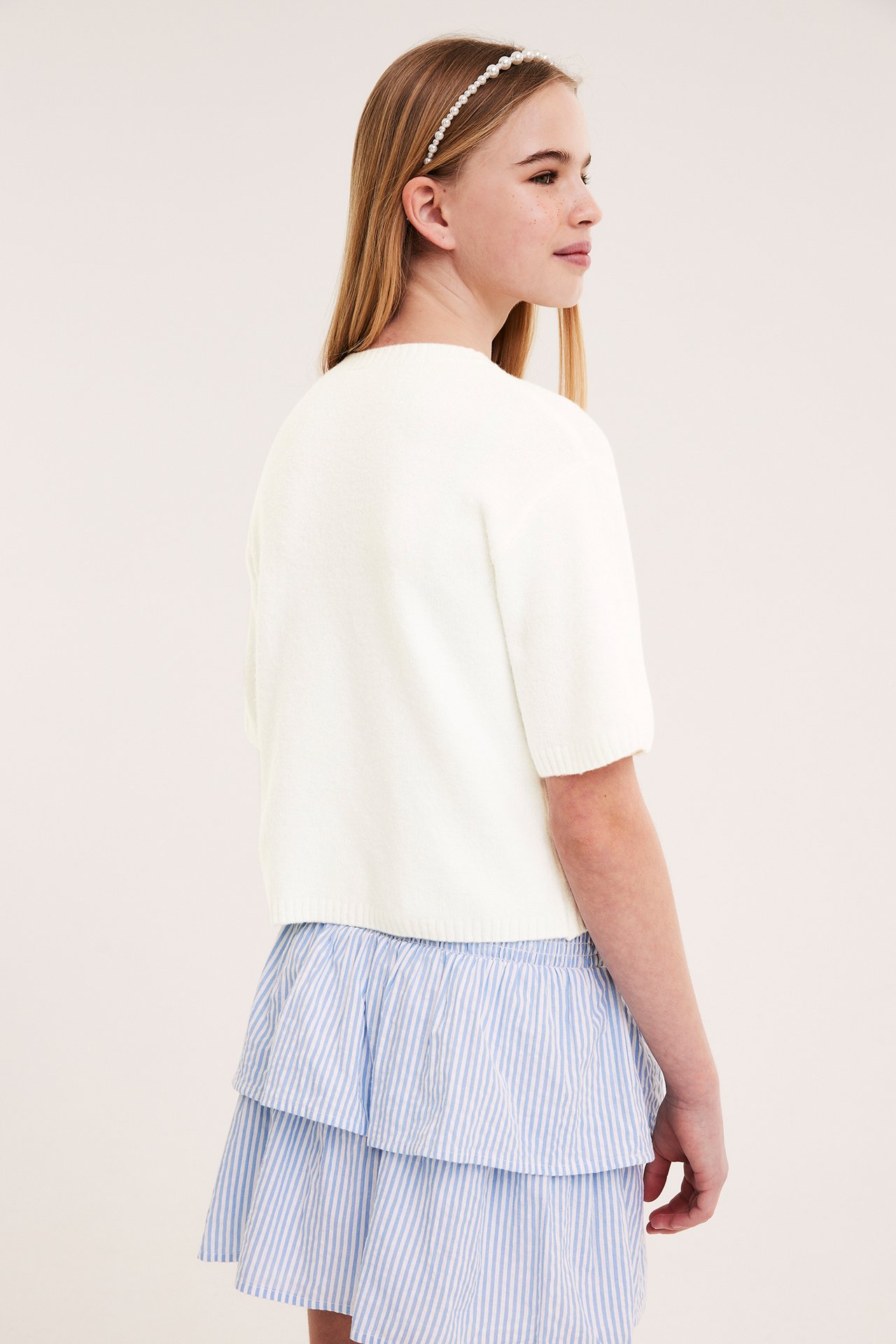 Widok z boku dziecka w ecru dzianinowym kardiganie z krótkim rękawem, niebieskiej spódnicy w paski i opasce z pereł.