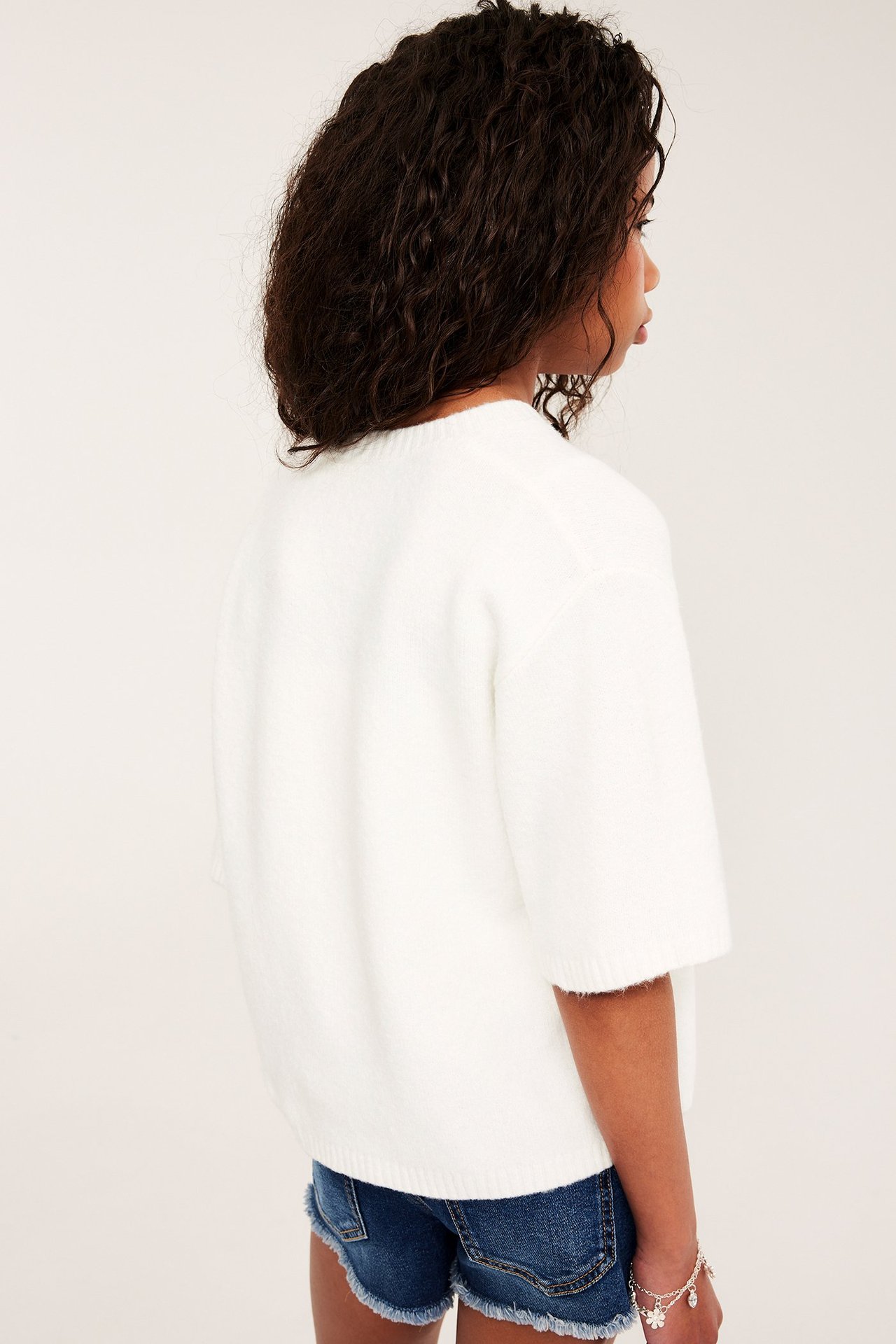 Bak-sidebilde av et barn i en offwhite kortermet strikket cardigan over blå denimshorts og et sølvarmbånd.