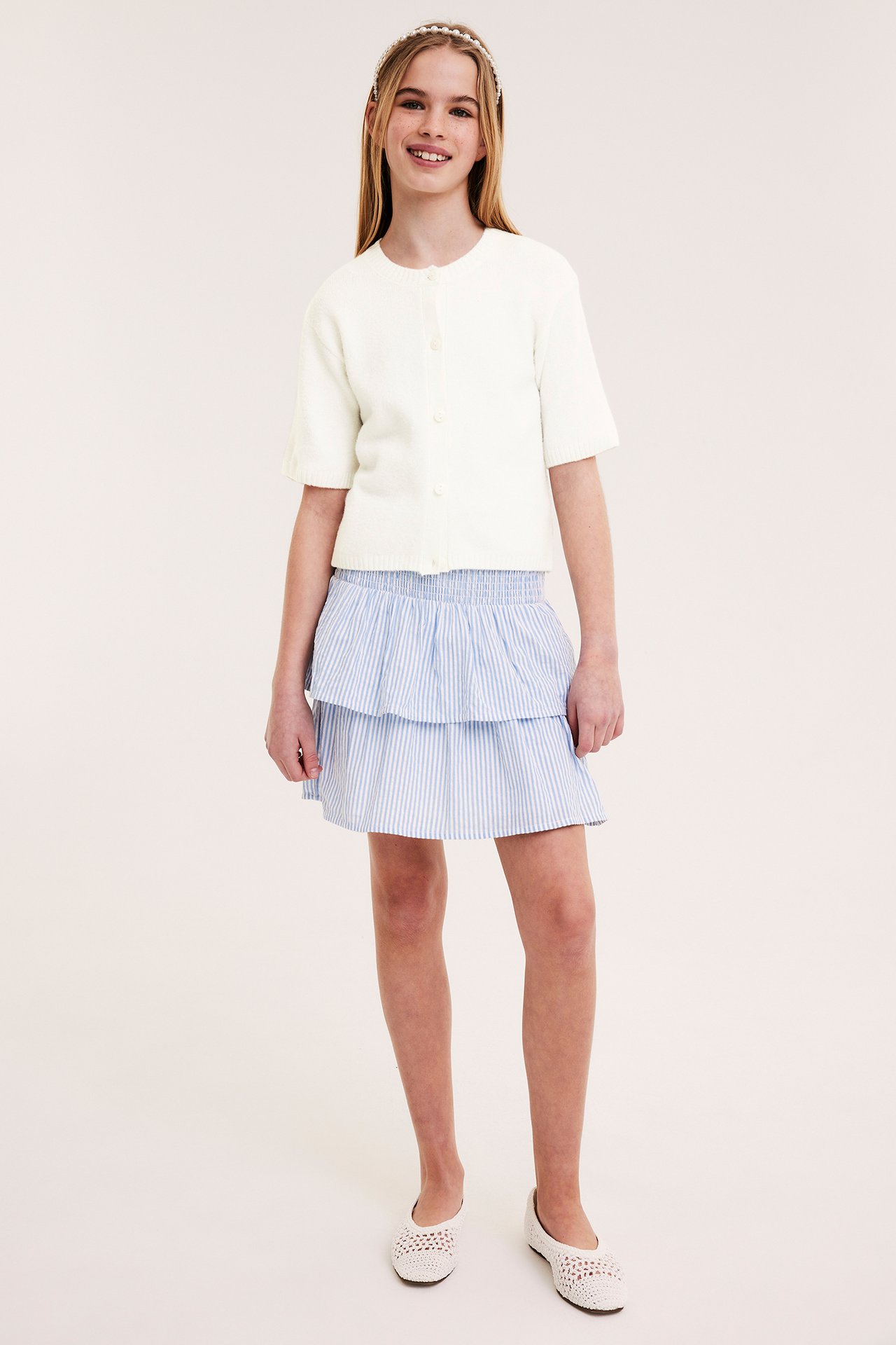 Framsida: Barn i offwhite kortärmad kofta, blårandig kjol, vita ballerinaskor och pärlhårband.