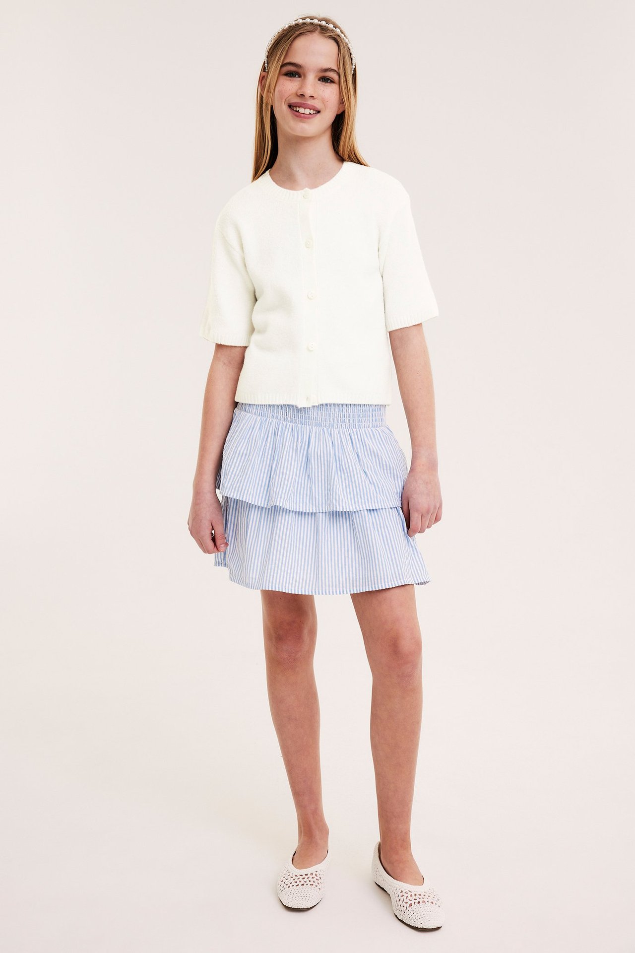 Frontvisning: Barn i offwhite kortermet cardigan, blåstripet skjørt, hvite ballerinasko og perlehårbånd.