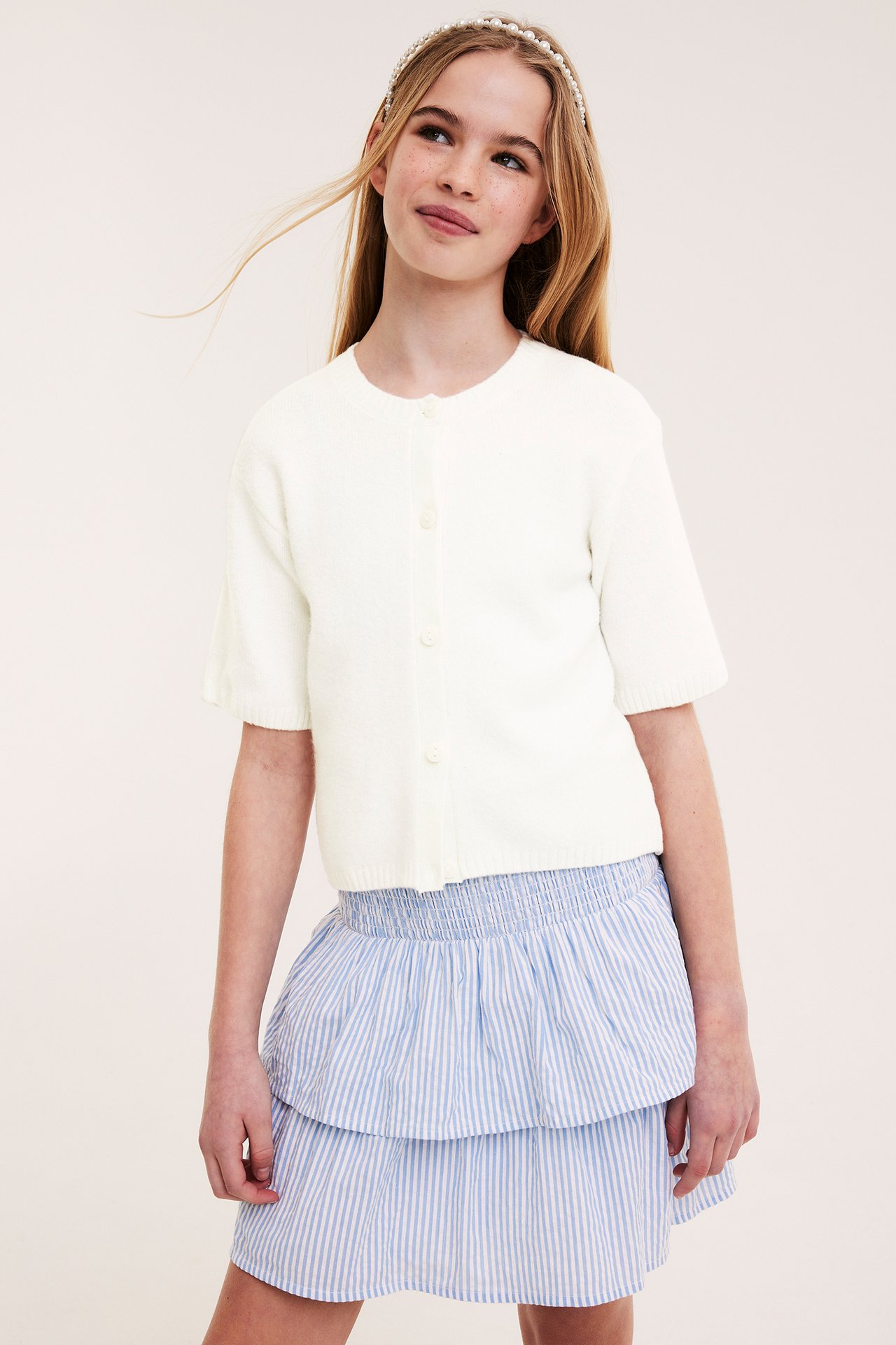 Dziewczynka w ecru dzianinowym kardiganie, niebieskiej spódnicy w paski i opasce z pereł.