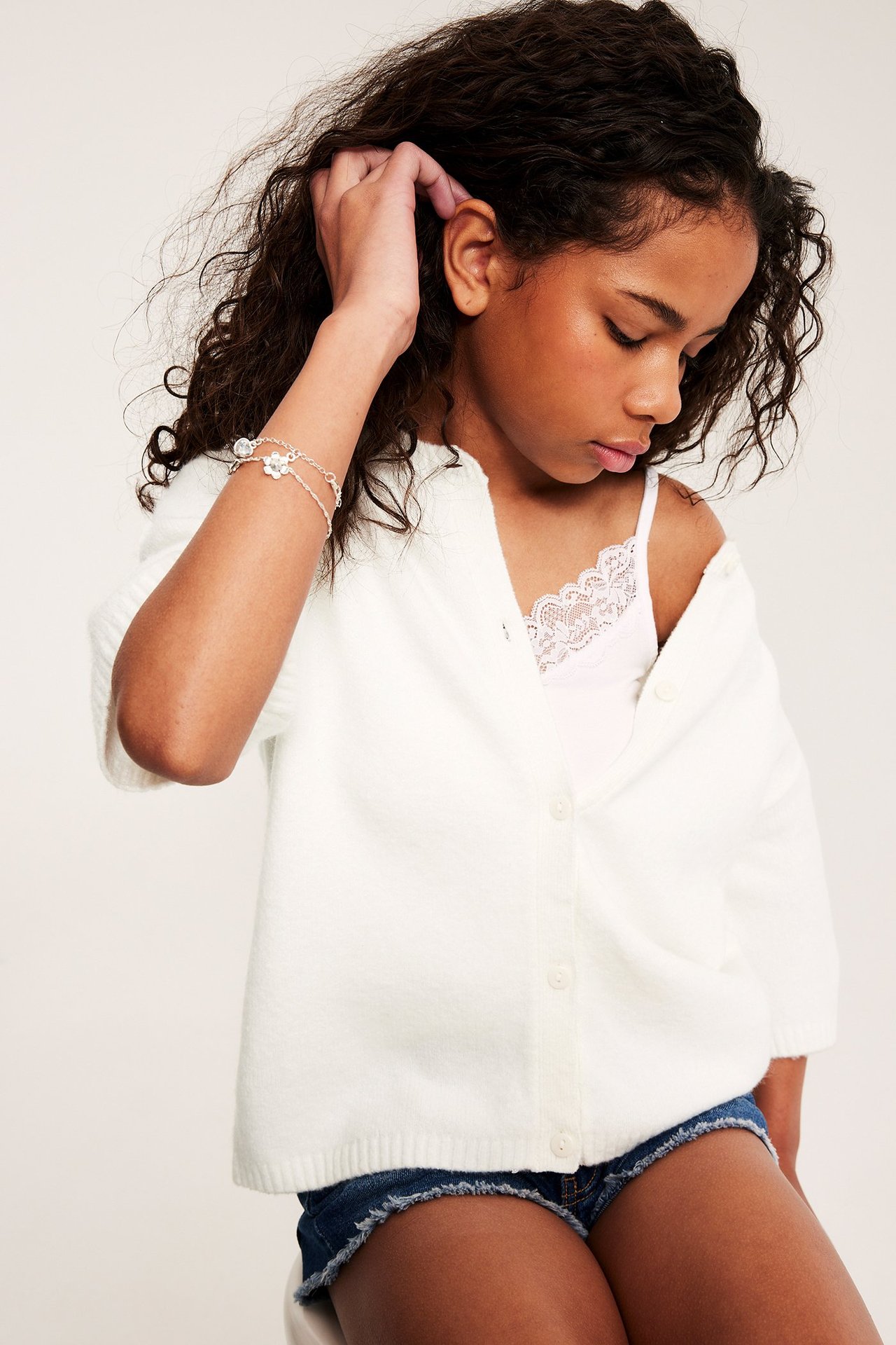 Edestäpäin: Lapsi pukeutuu luonnonvalkoiseen lyhythihaiseen neulejakkuun, valkoiseen pitsialuspaitaan, farkkushortseihin ja hopearannekoruun.