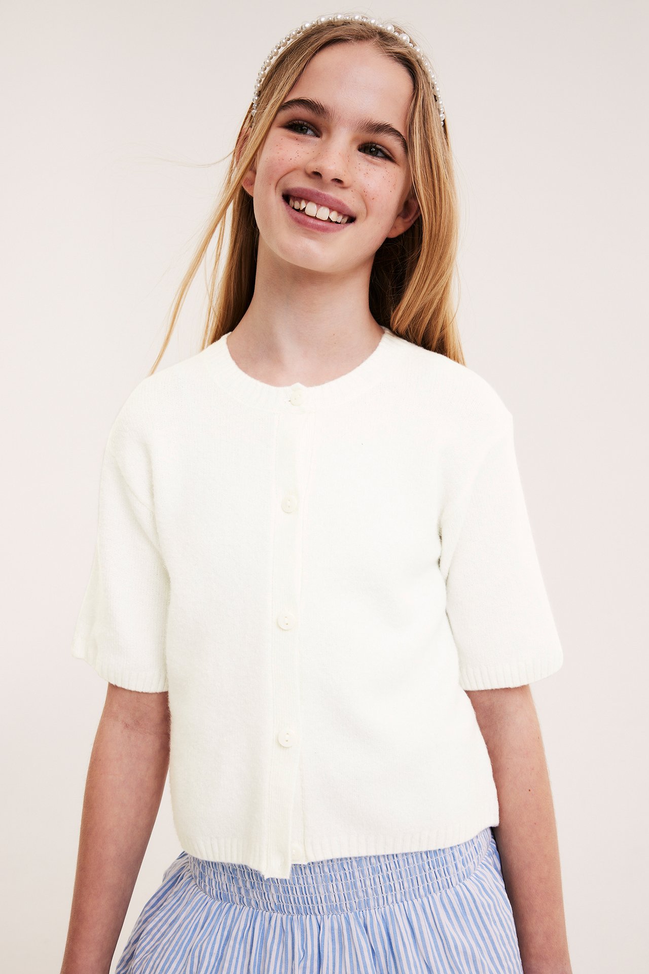 Framifrån: kid i en offwhite stickad kortärmad kofta, en blårandig kjol och ett pärlhårband.