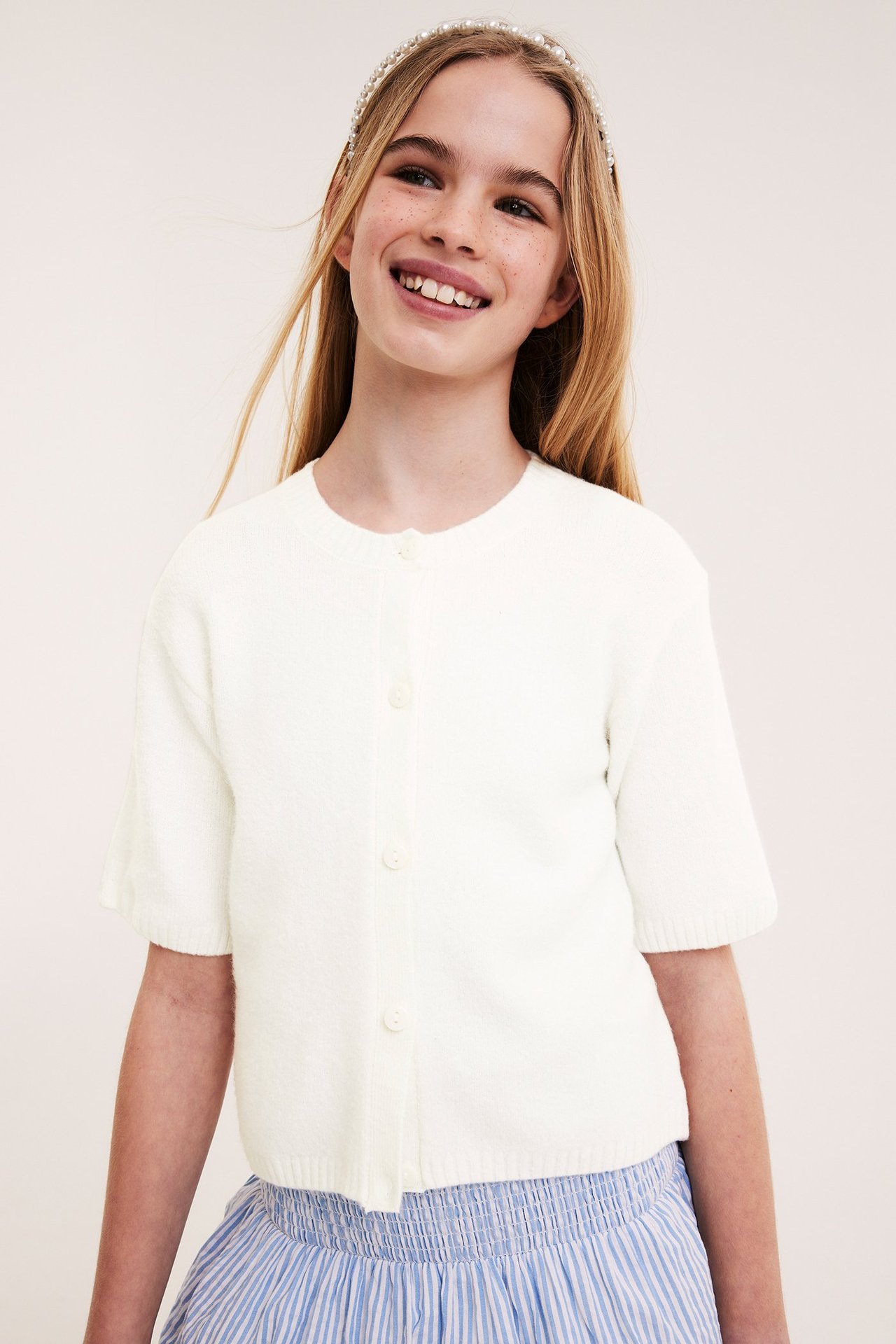 Forfra: barn i en offwhite strikket kortermet cardigan, et blåstripet skjørt og et perlehårbånd.