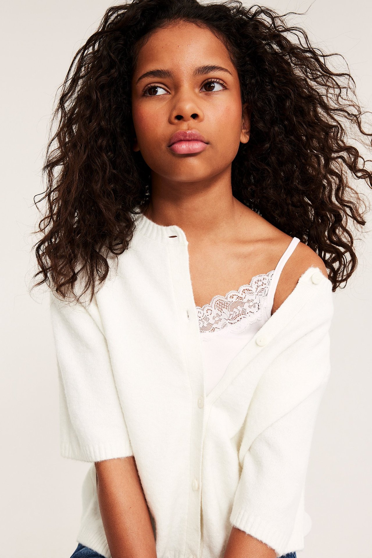 Nærbilde av et barn i en offwhite kortermet strikket cardigan over en hvit blonde-camisole.