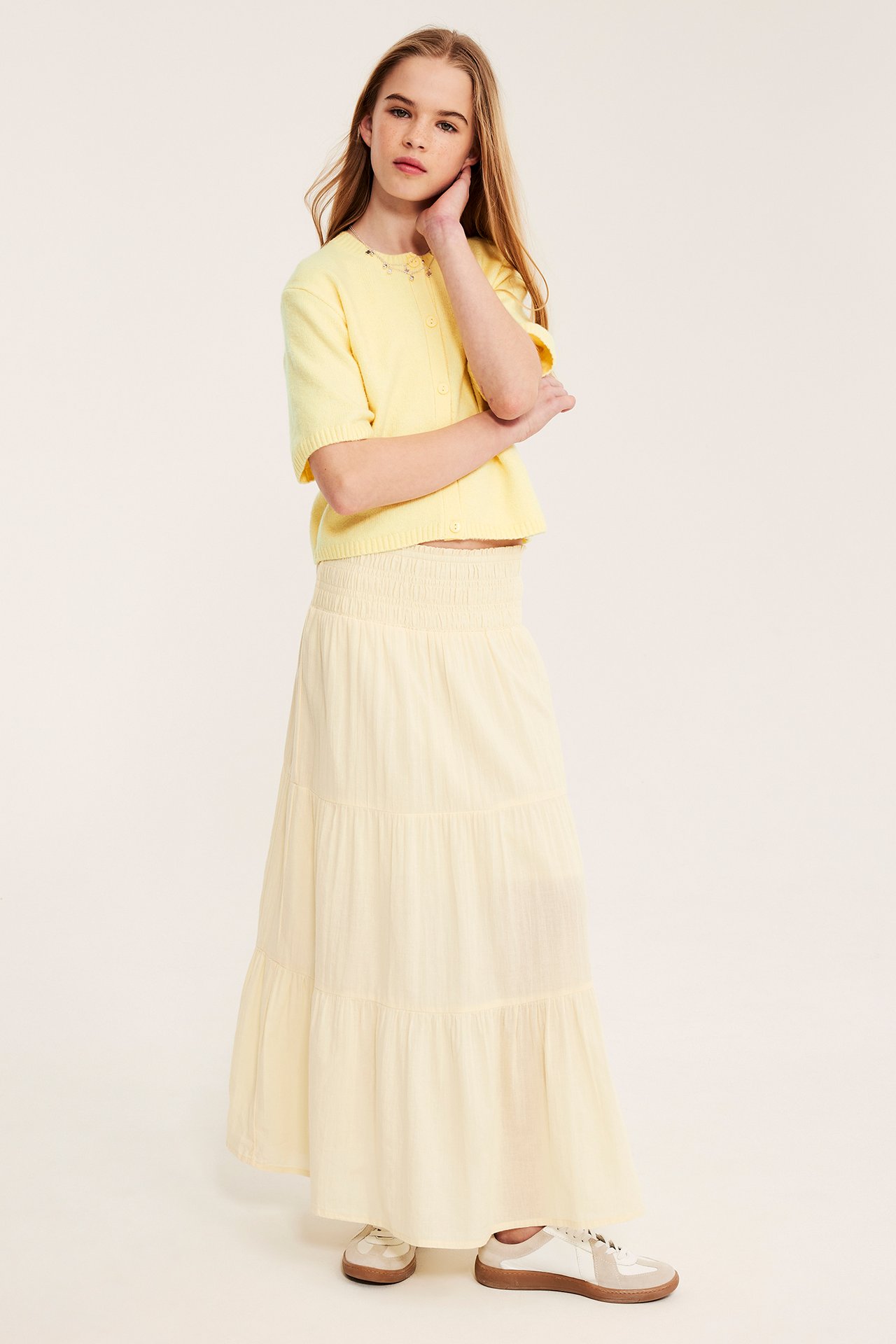 Frontbild: barn i ljusgul kortärmad stickad kofta med knappar, matchande maxikjol och sneakers.