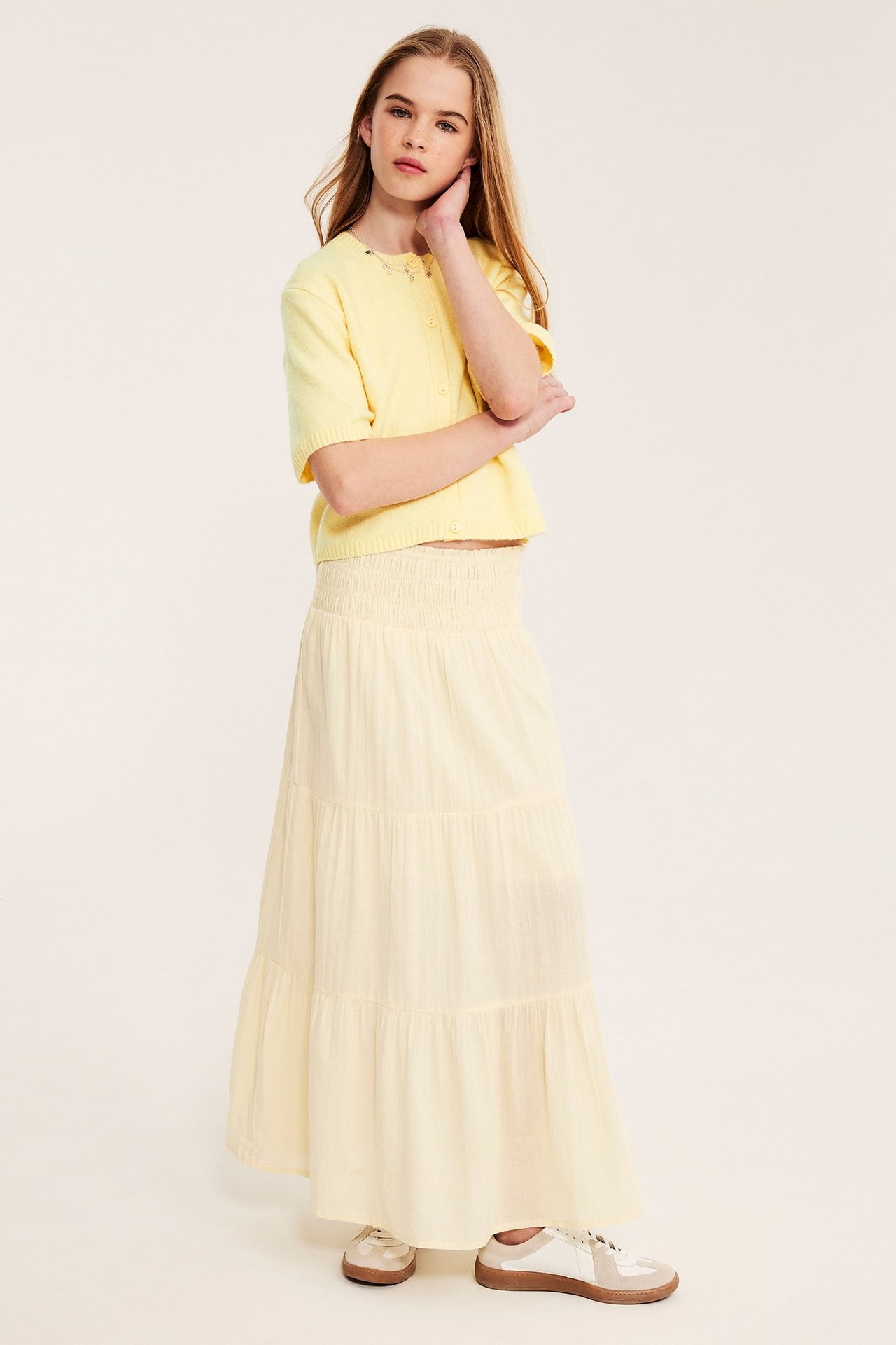 Frontbilde: barn i lysegul kortermet strikket cardigan med knapper, matchende maxiskjørt og joggesko.