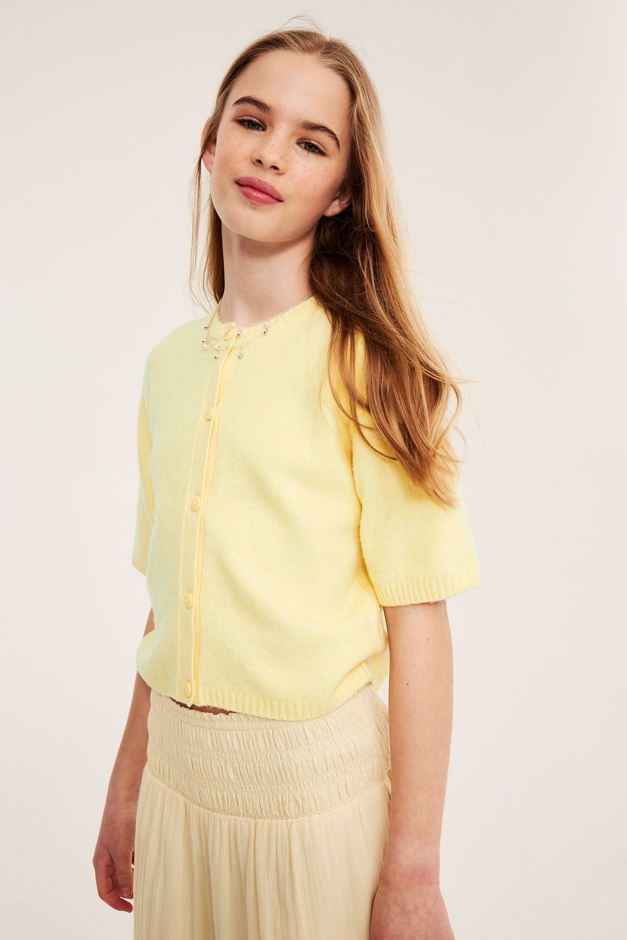 Frontvisning: Jente i lysegul kortermet strikket cardigan med knapper, lysebeige bukser og smykke.