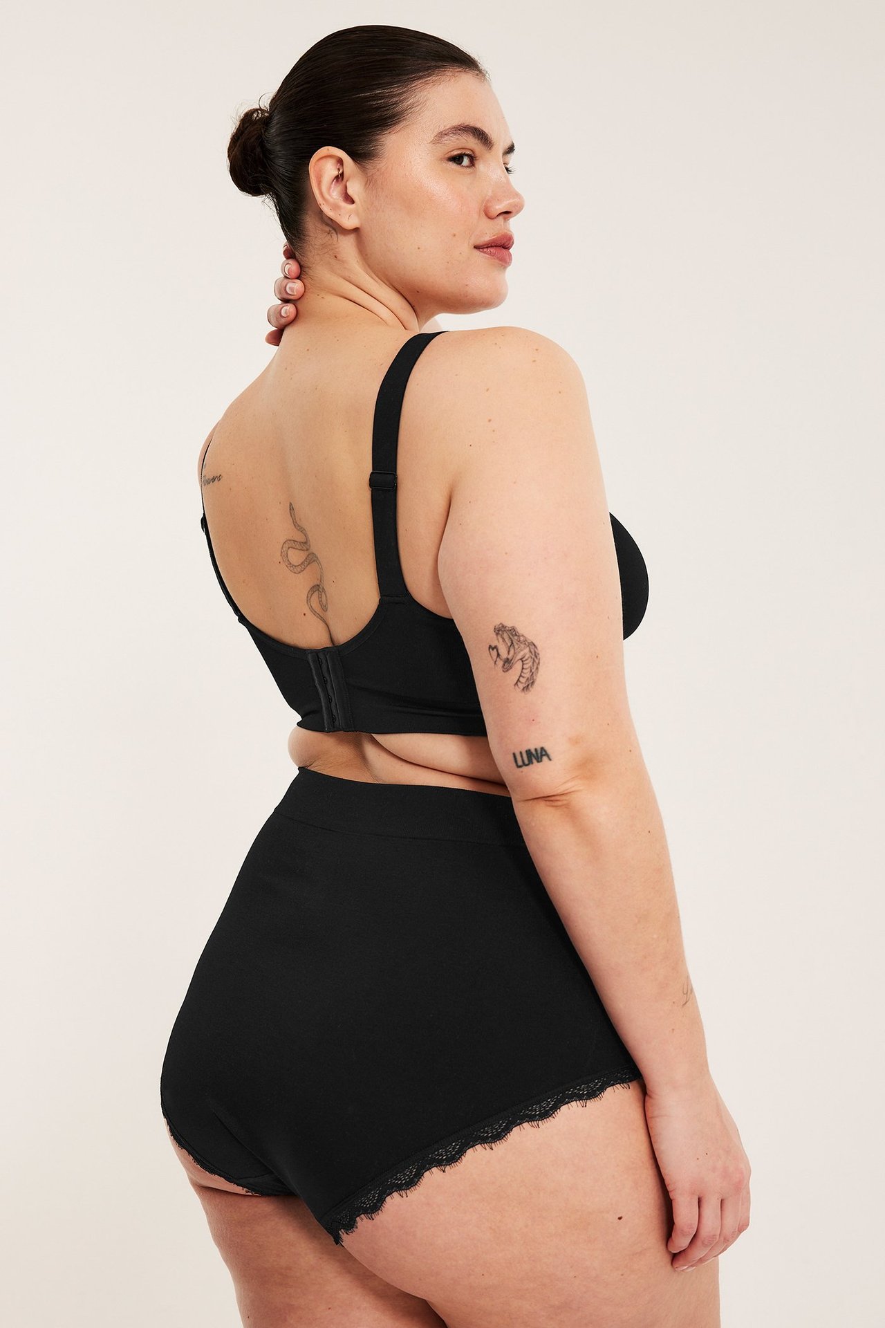 Nainen mustissa korkeavyötäröisissä hipstertrosissa pitsireunalla ja yhteensopivissa mustissa rintaliiveissä, takaa.