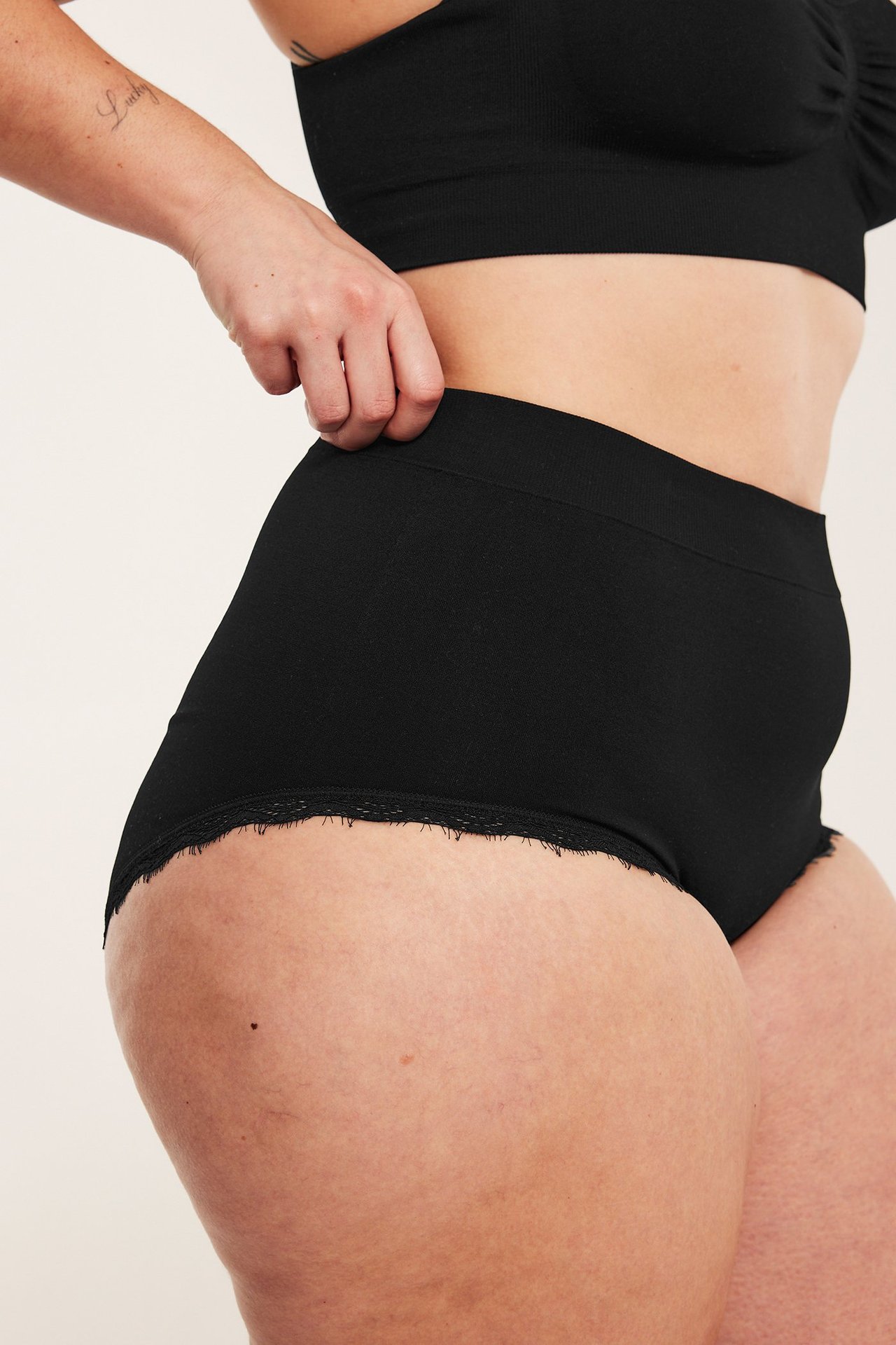 Sidevisning: kvinne i sorte sømløse hipster-truser med blondekant, trekker opp midjen, med ribbet BH.