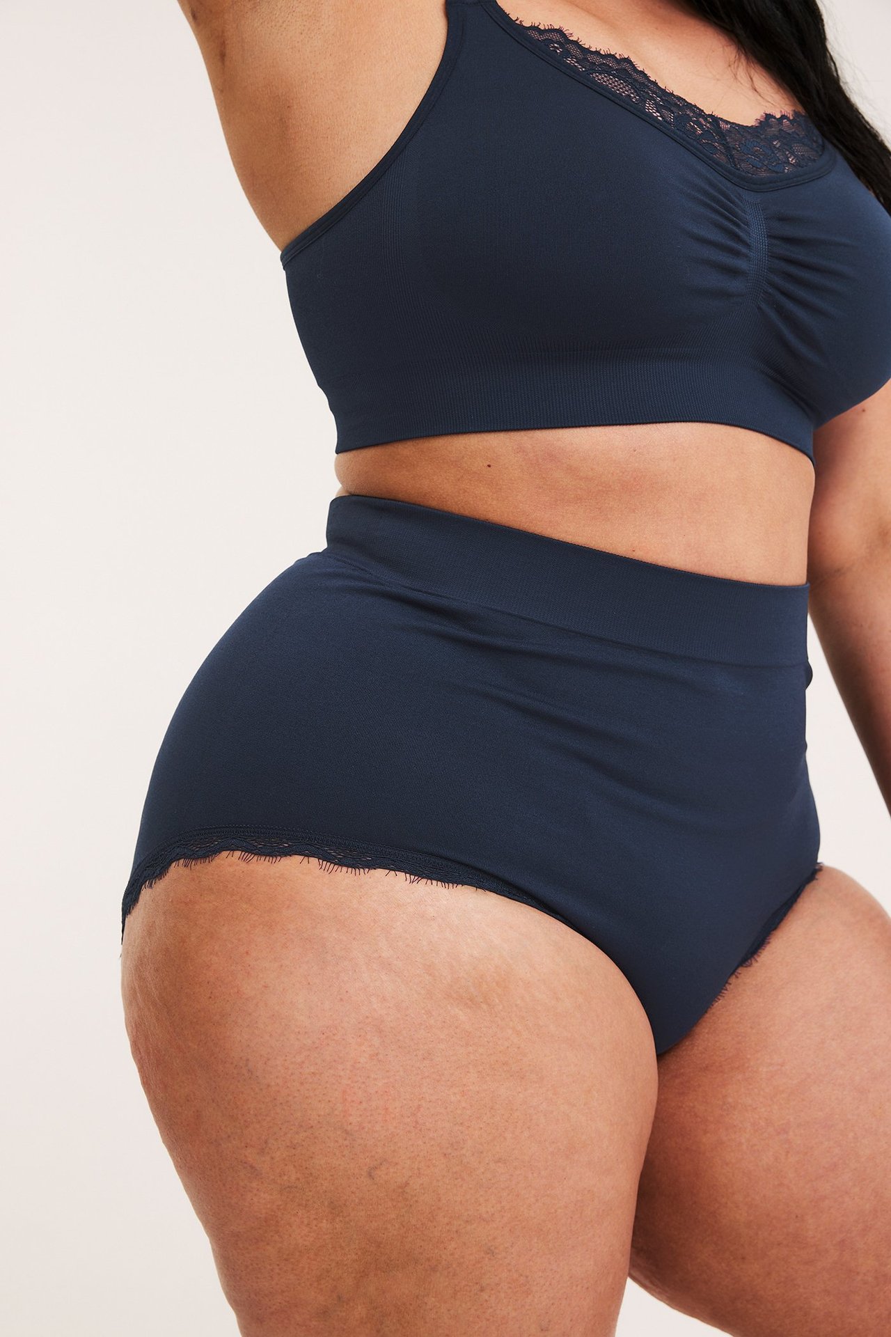Sidebilde av kvinne i mørkeblå sømløs høy hipstertruse med blondkant og matchende bralette.