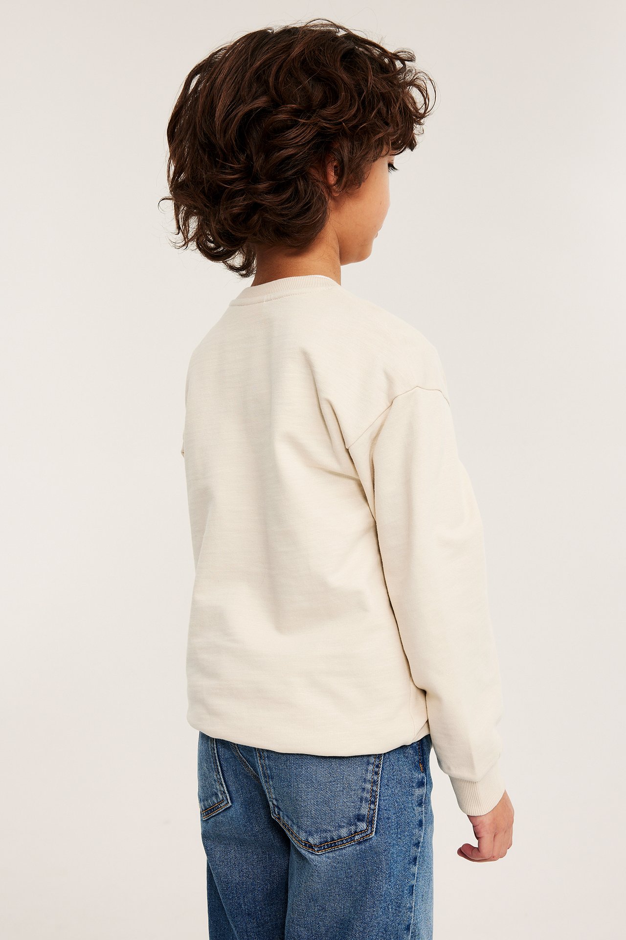 Lapsen taka-sivukuva, yllään oversized vaaleanbeige puuvillapaita ja siniset farkut.