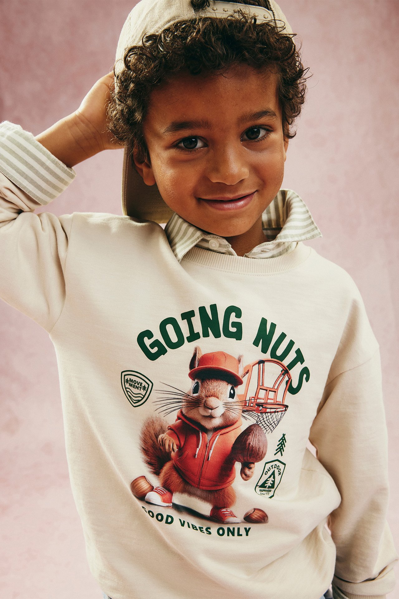 Frontvy på ett barn i ljusbeige sweatshirt med ekorrprint, randig skjorta och beige keps.