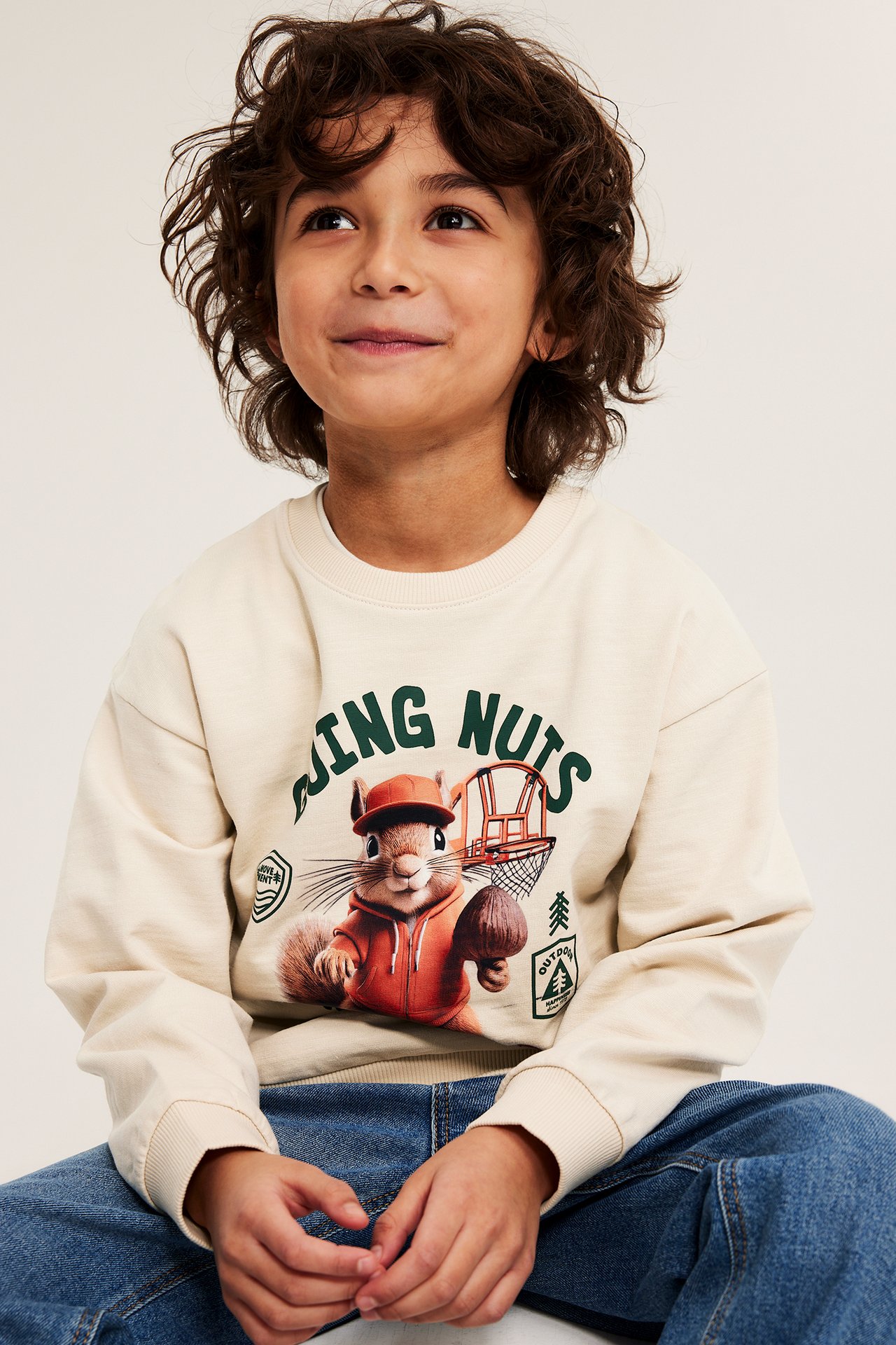 Frontvisning av et barn i lys beige, oversized bomullssweatshirt med ekorntrykk og blå jeans.