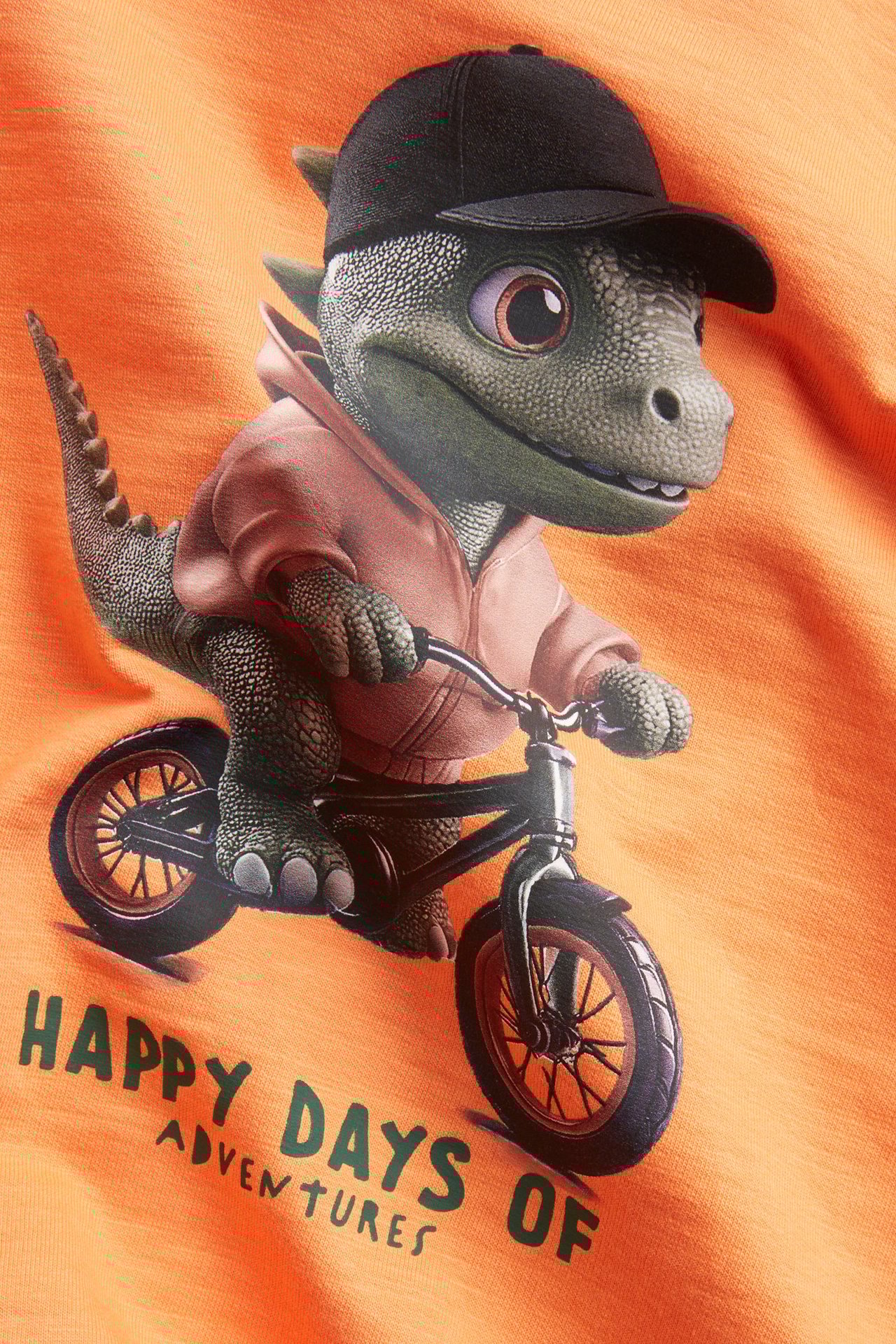 Lähikuva: Oranssi puuvillapaita, jossa dinosaurus mustassa lippiksessä ja hupparissa pyöräilee, ja teksti.