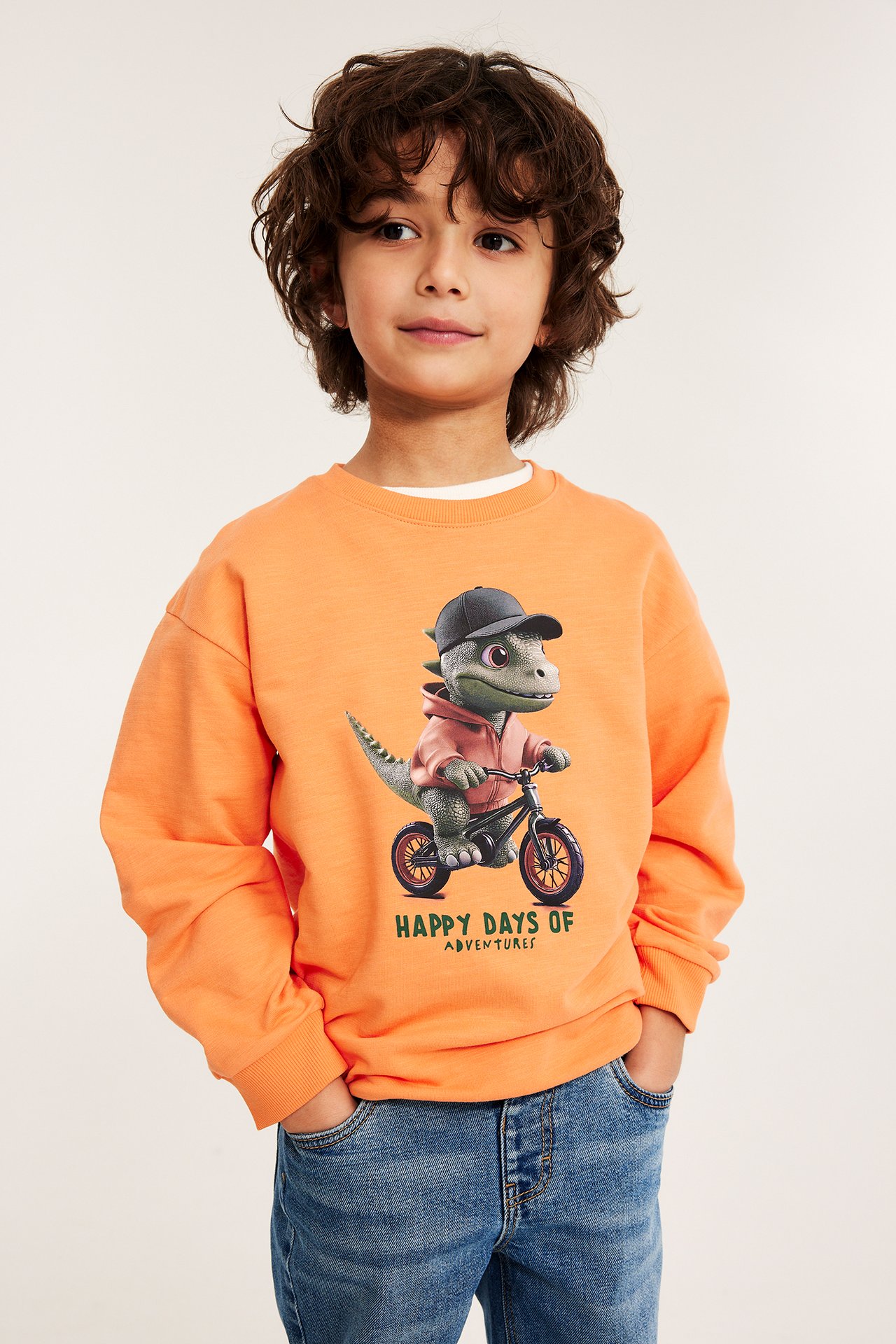 Forfra, et barn i en oransje oversized bomullssweatshirt med dinotrykk og blå jeans.