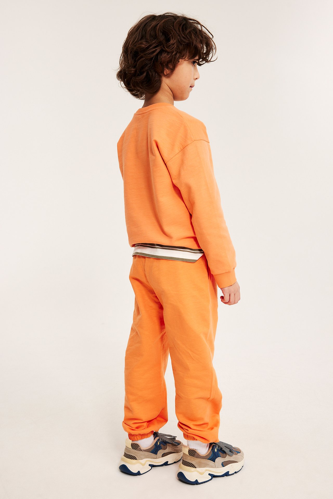 Sivukuva lapsesta, jolla on oranssit puuvillahousut painatuksella, oranssi collegepaita ja lenkkarit.