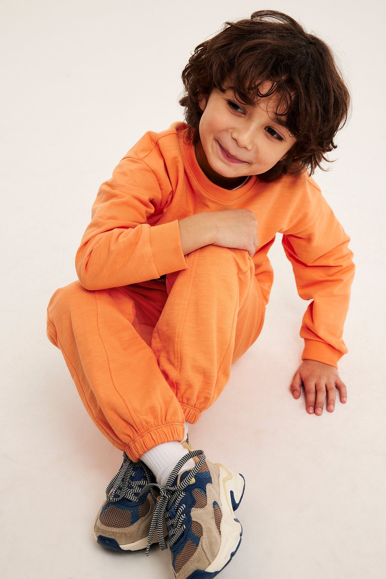 Edestä: Lapsi oransseissa puuvillacollegehousuissa, yhteensopivassa paidassa, valkoisissa sukissa ja lenkkareissa.