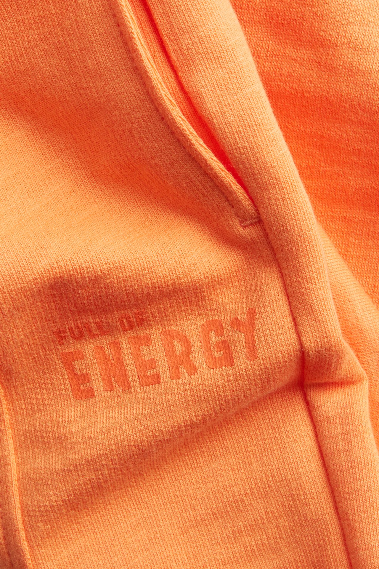 Närbild på orange bomulls joggingbyxor med en framficka och texttrycket 'FULL OF ENERGY'.