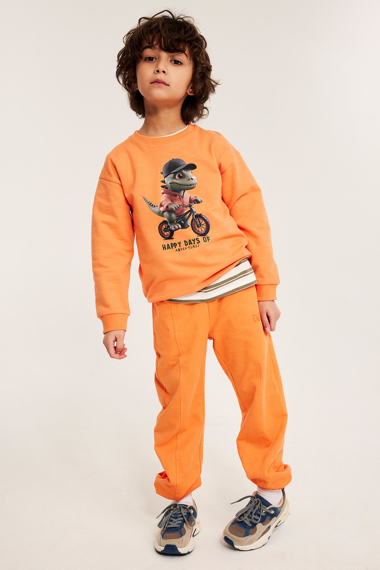 Frontvisning: Barn i oransje myk bomullsjoggebukse med trykk, stylet med en oransje dinosaur-collegegenser.
