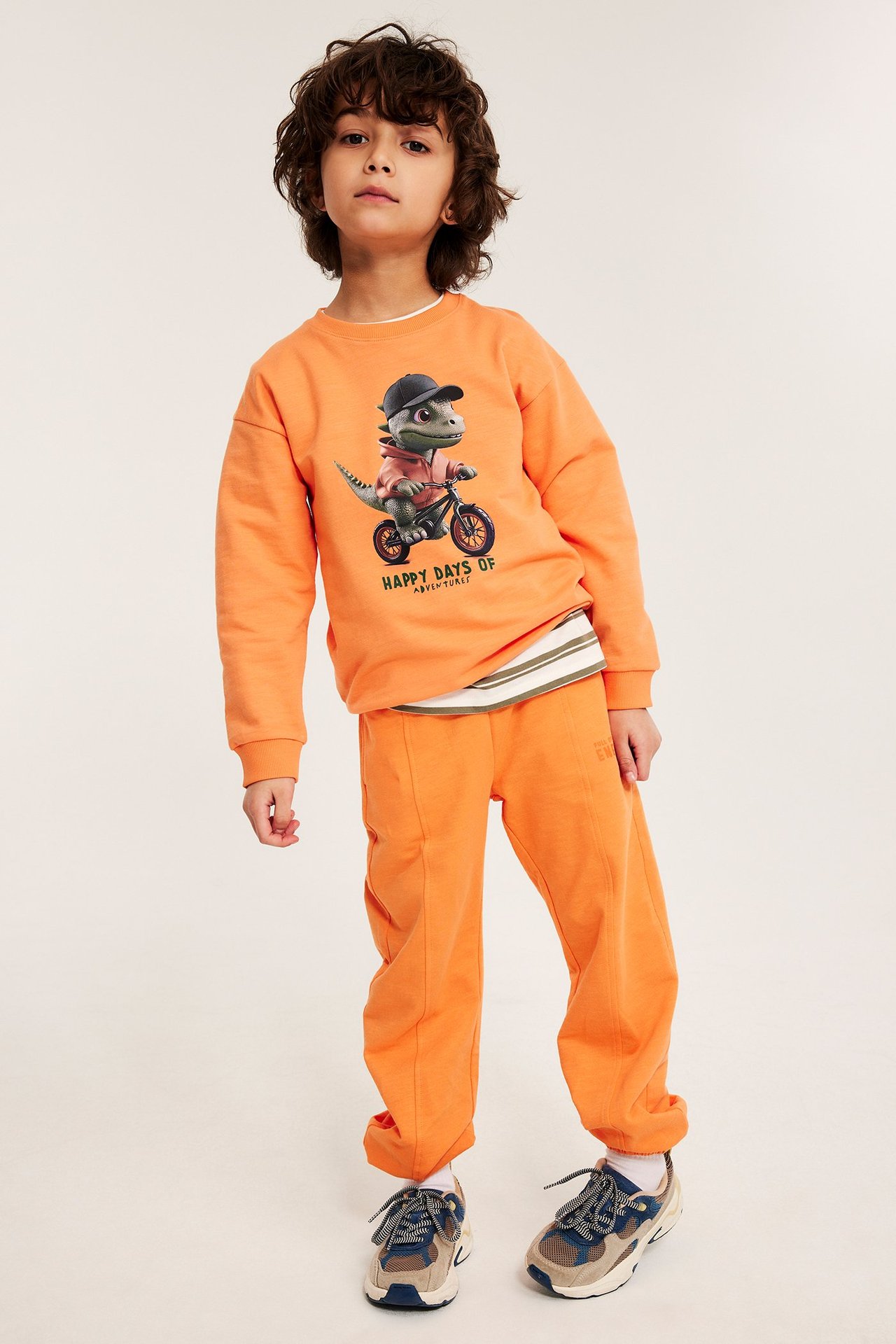 Etunäkymä: Lapsi oransseissa pehmeissä puuvillahousuissa, joissa tekstituloste, yhdistettynä oranssiin dinosauruscollegepaitaan.