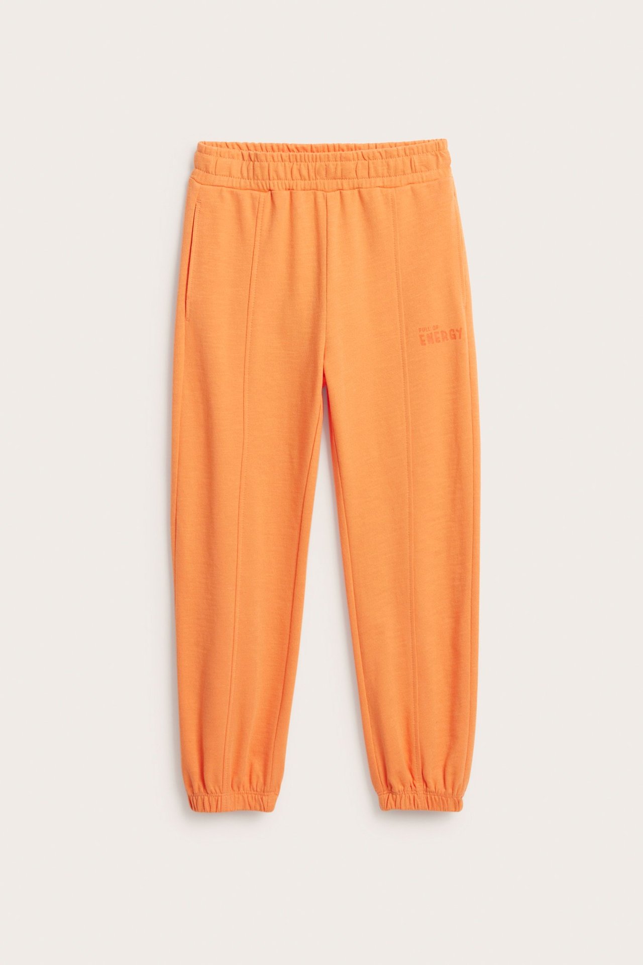 Framifrån: Orangea mjukisbyxor i bomull med resår i midja, fickor, bensömmar, muddar och lårprint.