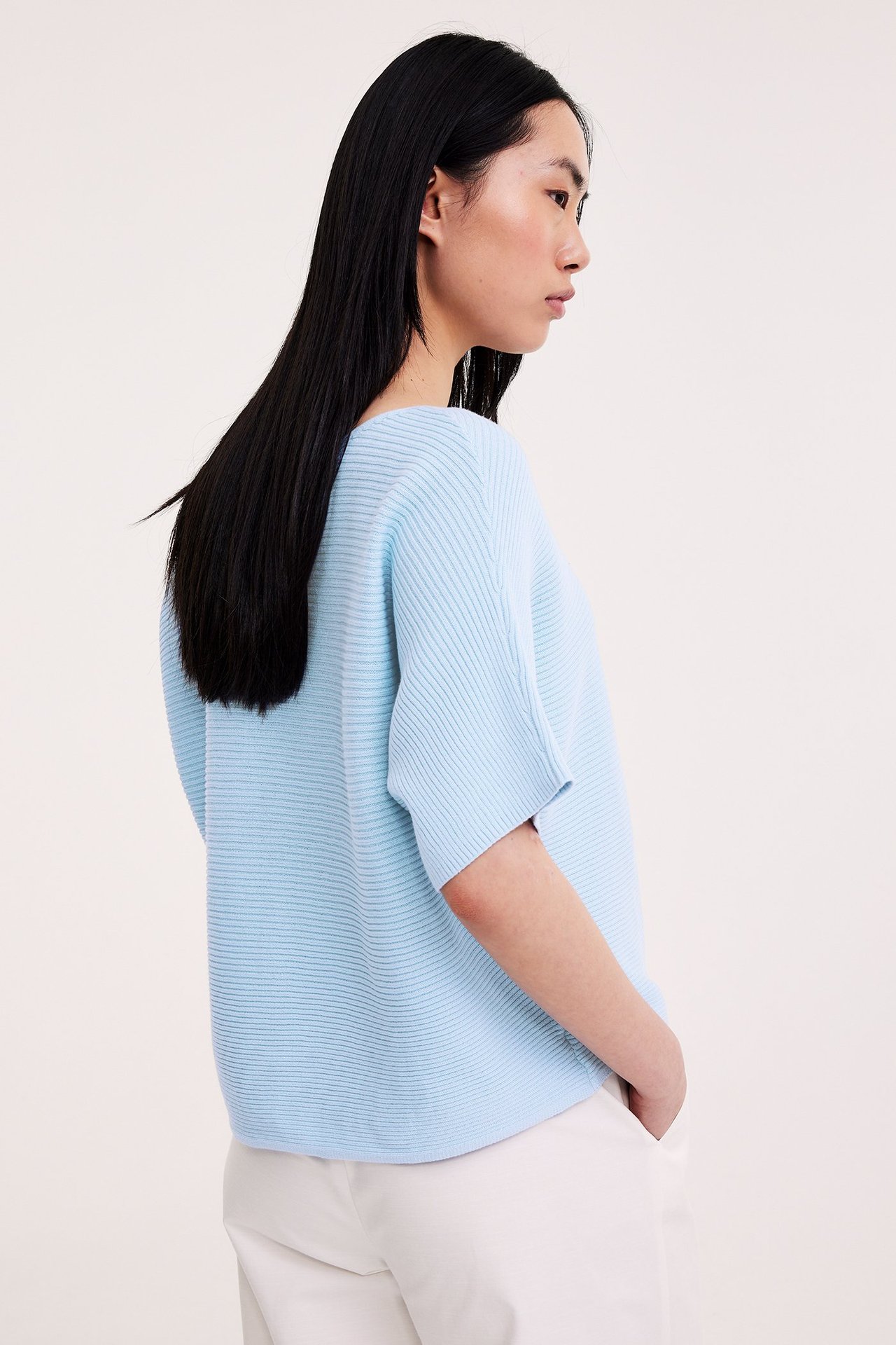 Sivukuva naisesta vaaleansinisessä, lyhythihaisessa, ribbineulotussa puuvillatopissa ja valkoisissa housuissa.