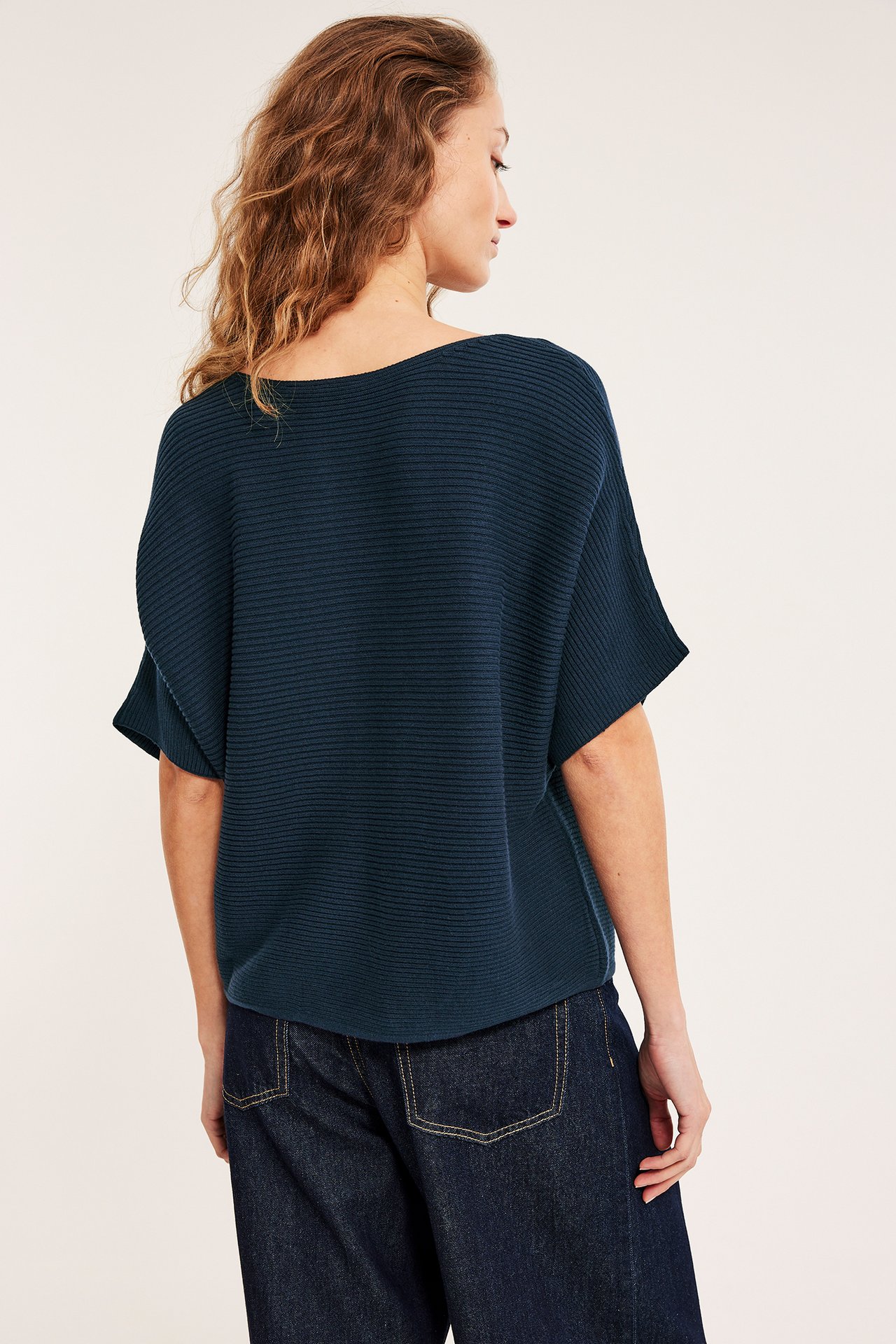 Bakfra: kvinne i mørkeblå ribbestrikket bomullsgenser, korte ermer, og mørkeblå vide jeans.