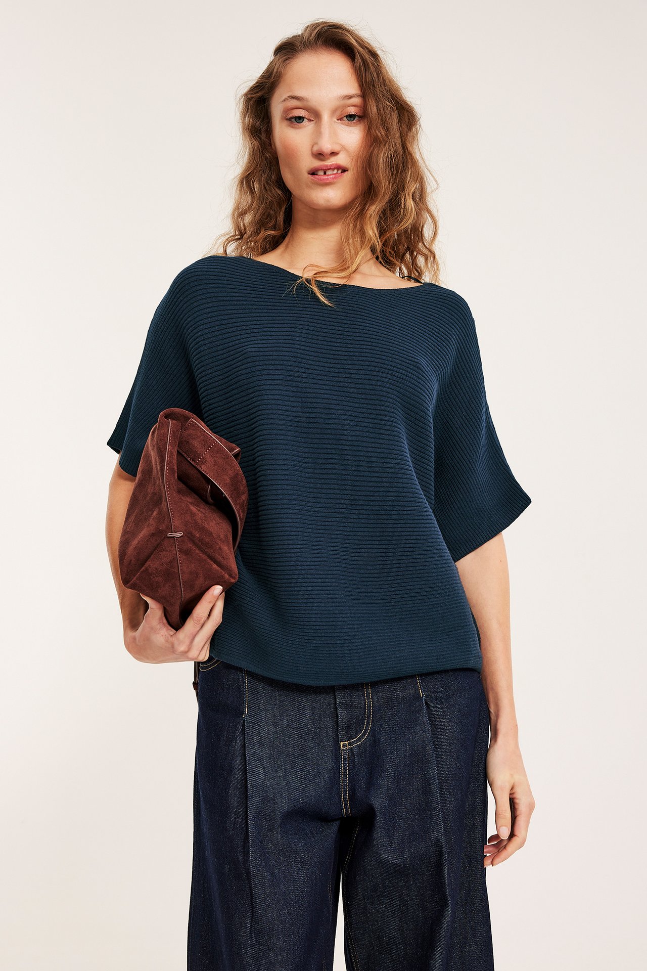 Frontbilde: Kvinne i mørkeblå ribbestrikket båthalsgenser, mørke jeans, holder en brun clutch.