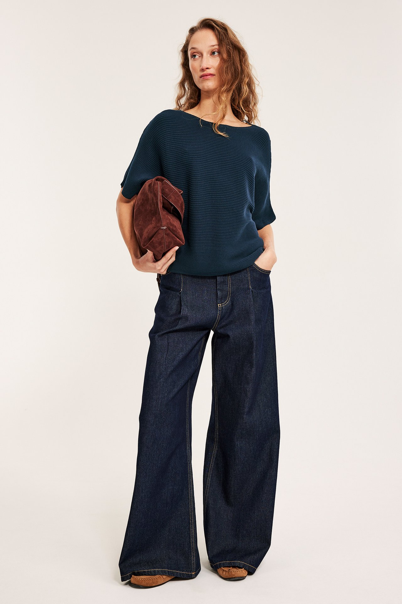 Forfra: Kvinne i mørkeblå ribbestrikket kortermet genser, vide jeans, holder en brun semsket clutch.
