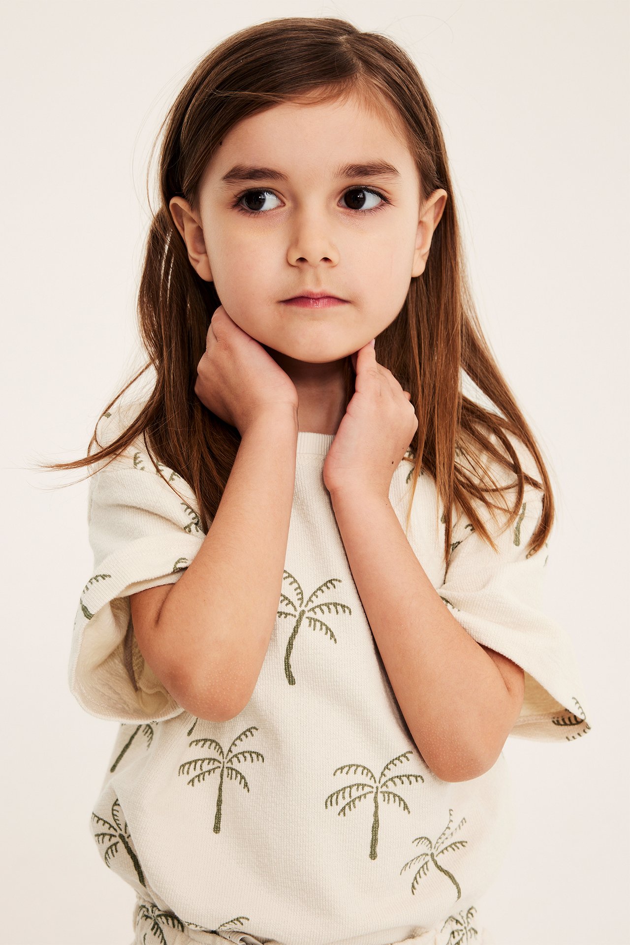 Forfra, et barn i en lys beige, romslig bomulls-T-skjorte med palmemønster.