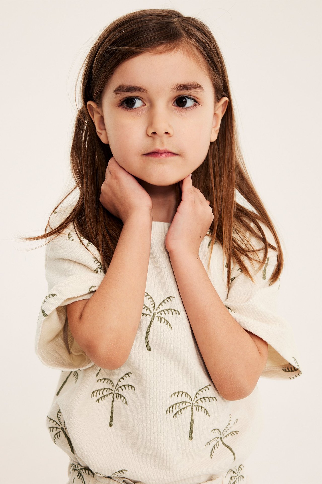Edestä kuvattu lapsi yllään vaaleabeige, väljä puuvillainen t-paita palmupuukuviolla.