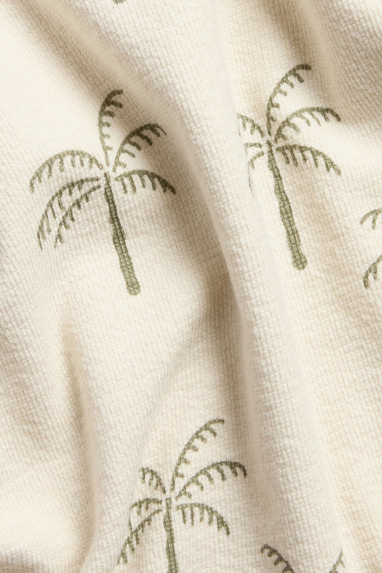 Zbliżenie na jasnobeżowy materiał koszulki z krepy bawełnianej z całopowierzchniowym wzorem zielonych palm.