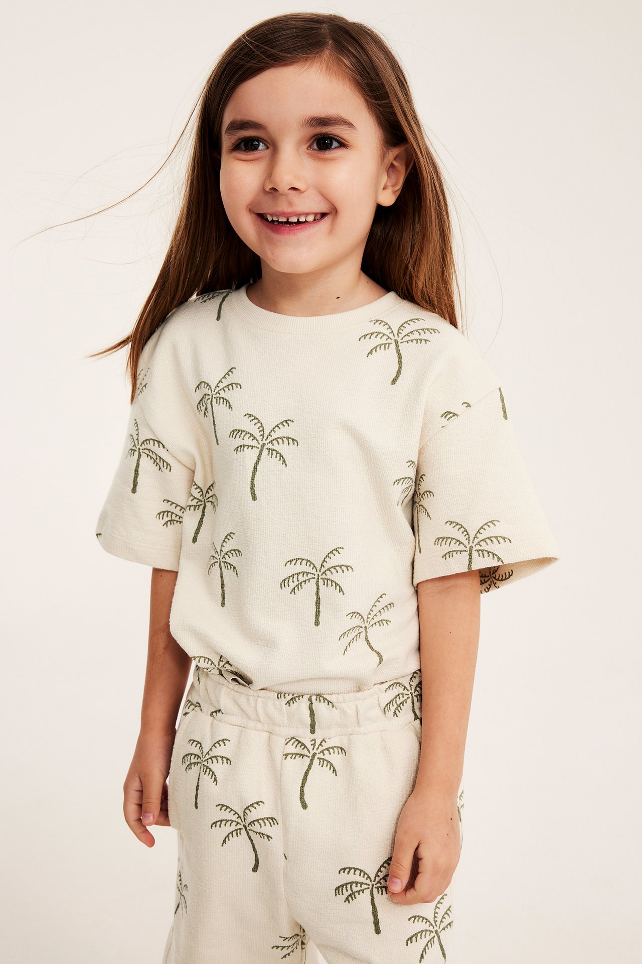 Frontutsikt: Barn i løs lys beige bomullskrep-t-skjorte og matchende shorts med palmeprint.