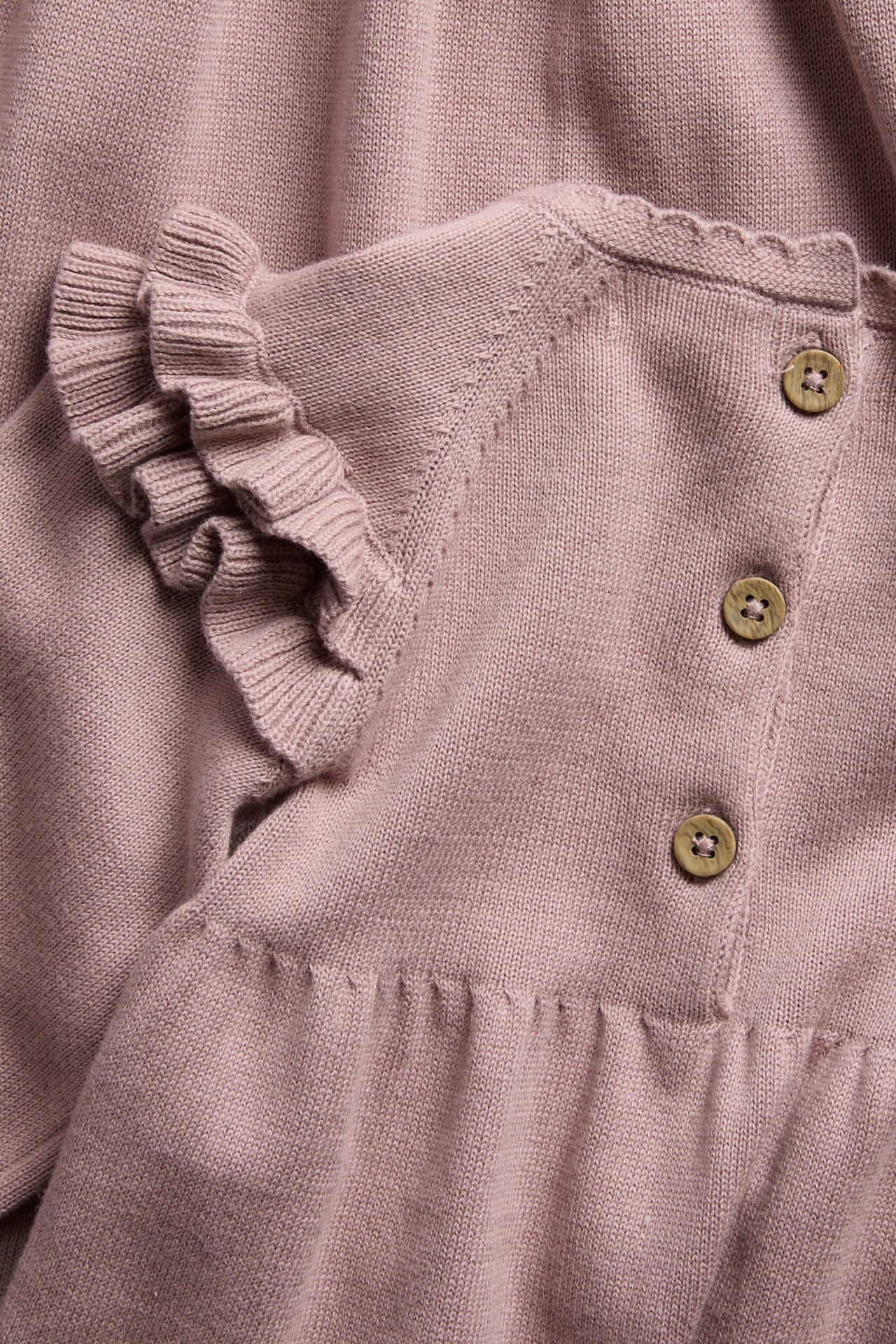 Närbild av ett rosa stickat babyplagg med volangärmar, träknappar och en vågig kant.