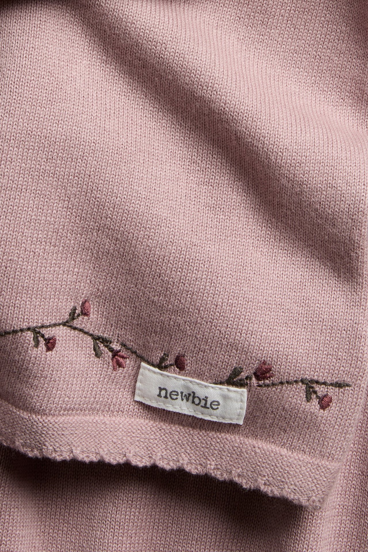 Närbild på ett dammrosa stickat babyplagg med broderad blomdetalj och vit etikett.