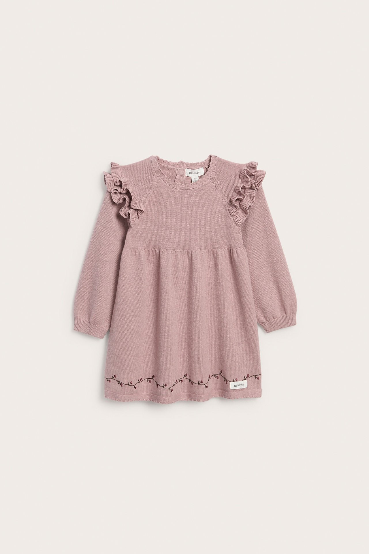 Framifrån, en babyrosa stickad klänning med volanger på axlarna och broderade vinstockar.