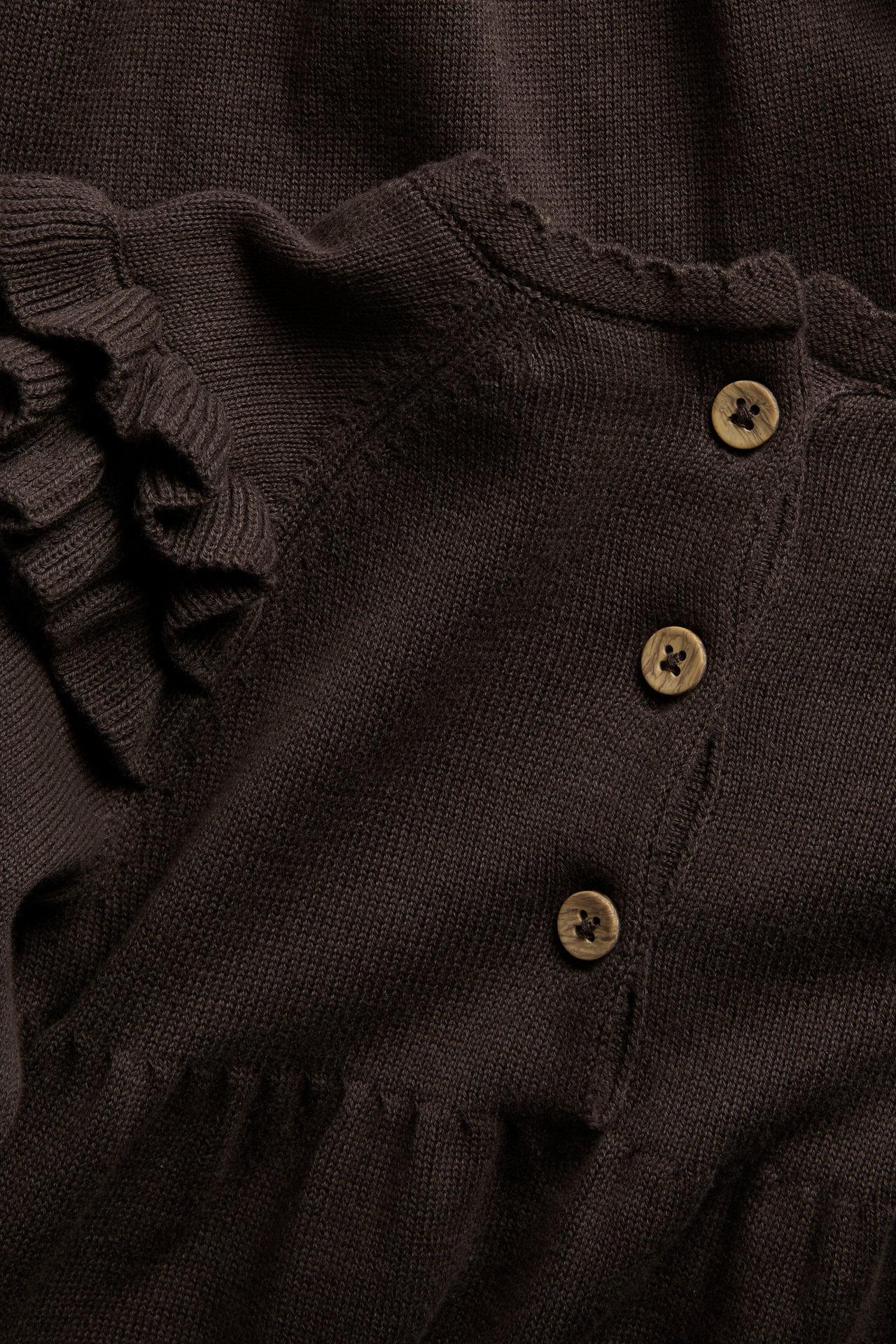 Närbild av ett mörkbrunt stickat plagg med vågig kant, volangärm och träknappar.
