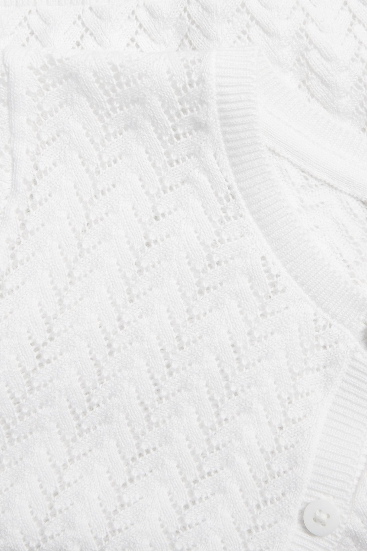 Närbild av en vit mönsterstickad bomullskofta med ribbad kant och en vit knapp.