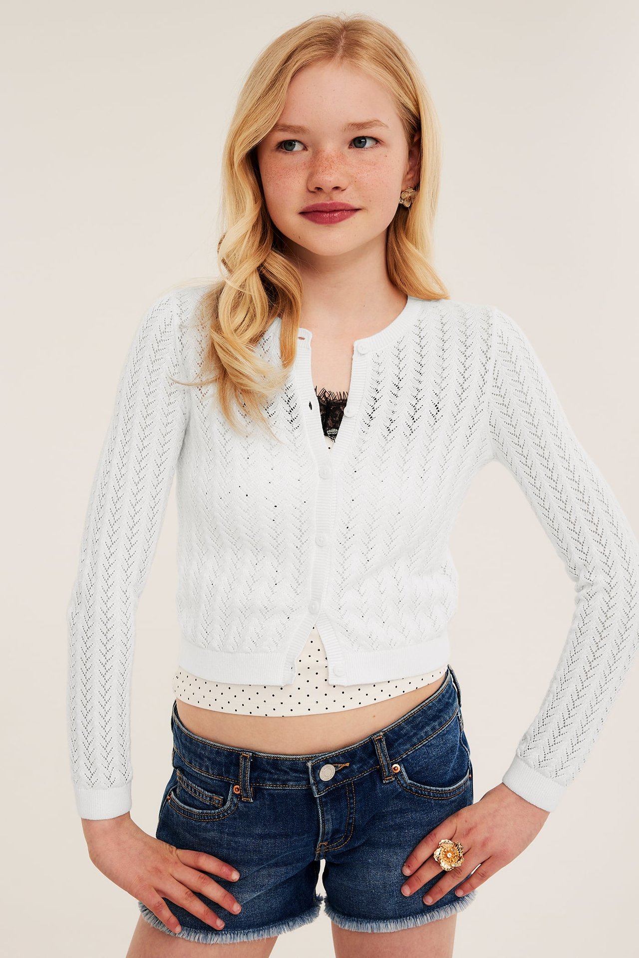 Front: Barn i hvit mønsterstrikket cardigan, over svart topp og blå denimshorts.