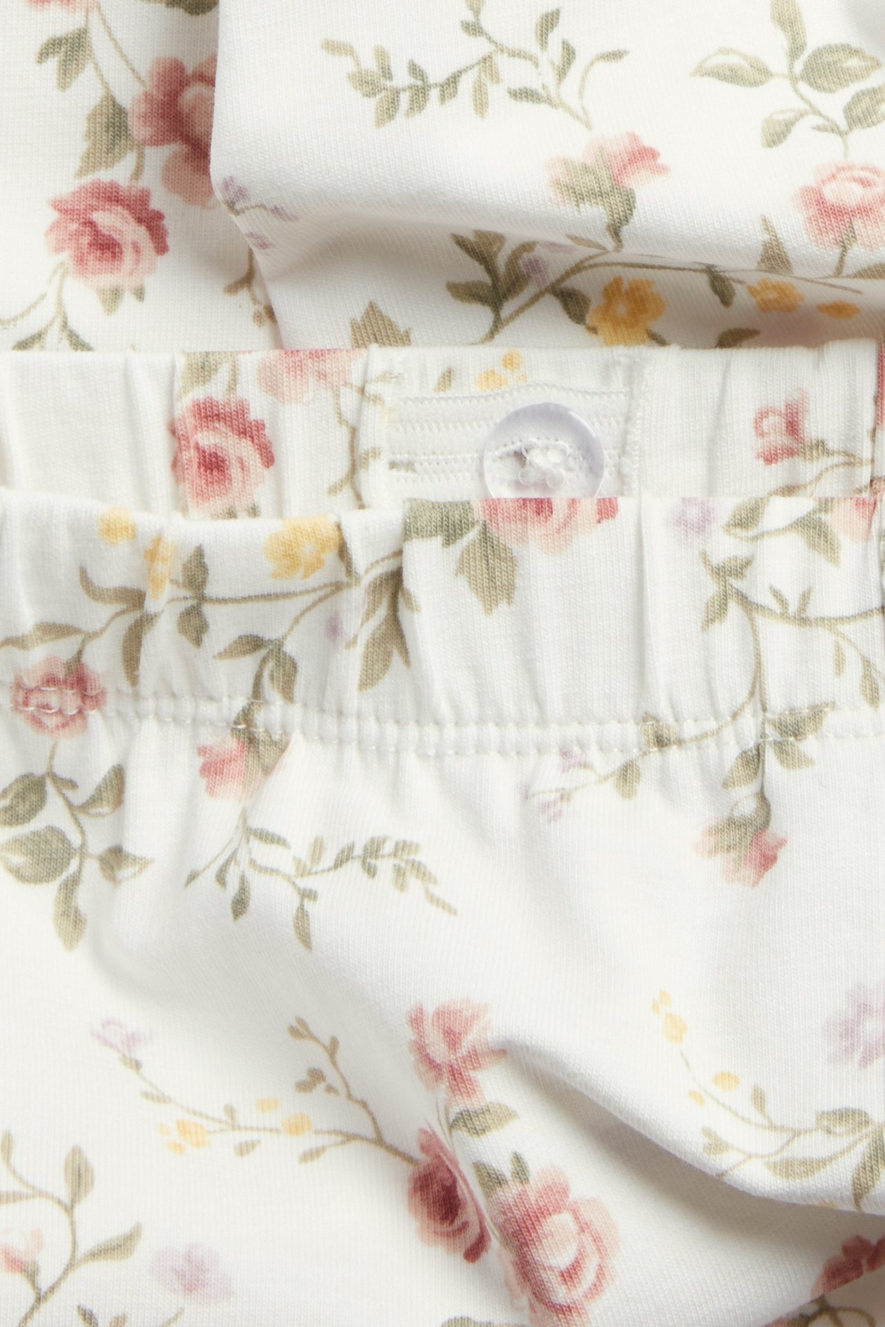 Nærbilde av offwhite blomstrete leggings med elastisk linning og gjennomsiktig knapp.