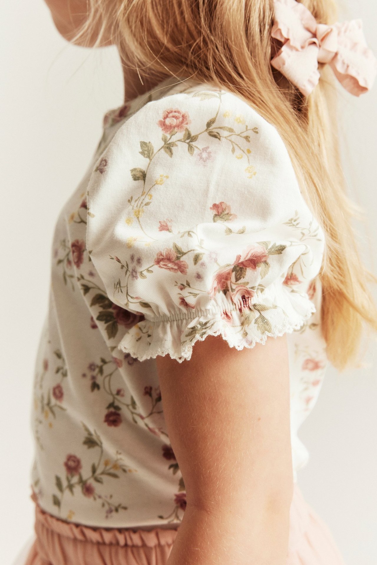 Sidevisning: Barn i offwhite blomstret jerseytop med puffermer, rosa skjørt og rosa hårsløyfe.