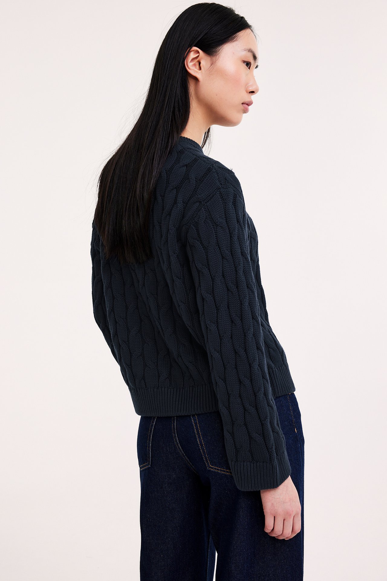 Widok z boku kobiety w granatowym bawełnianym swetrze z warkoczami i granatowych jeansach.