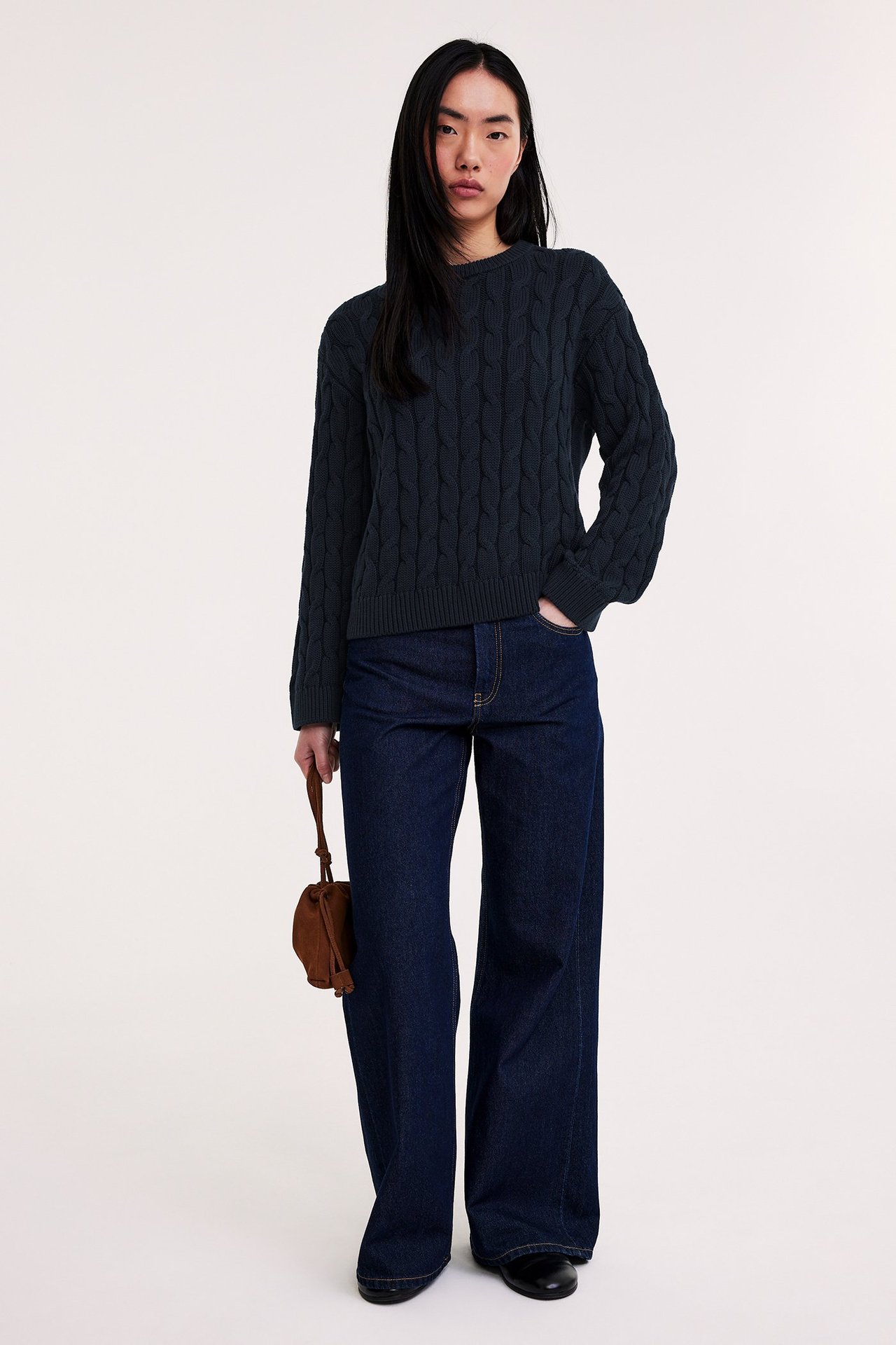 Framifrån: kvinna i mörkblå kabelstickad bomullströja, mörkblå jeans, håller brun mockaväska.
