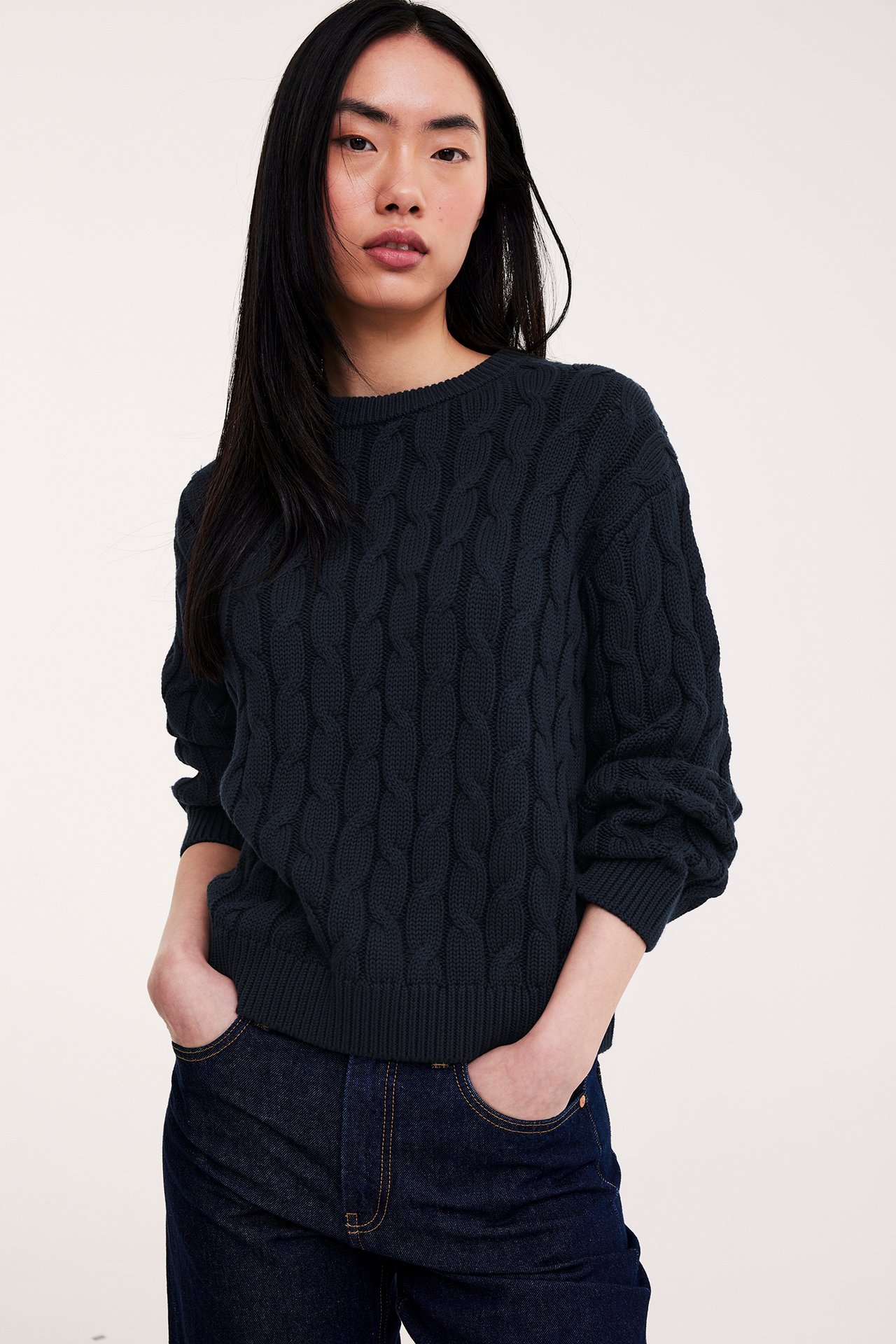 Framifrån: kvinna i mörkblå kabelstickad bomullströja och mörkblå jeans, med händerna i fickorna.