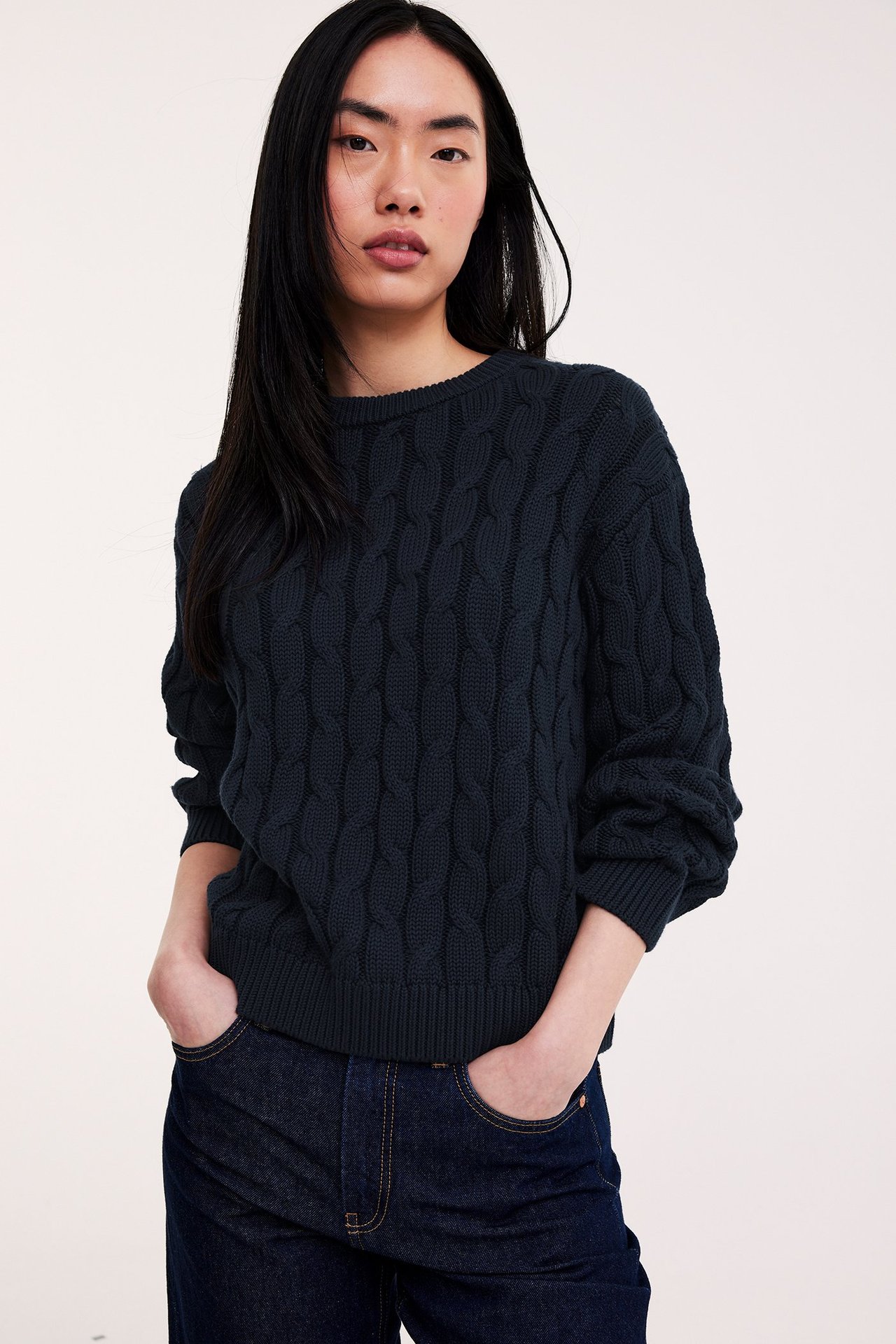 Kobieta w granatowym, bawełnianym swetrze z warkoczowym splotem i jeansach, ręce w kieszeniach.