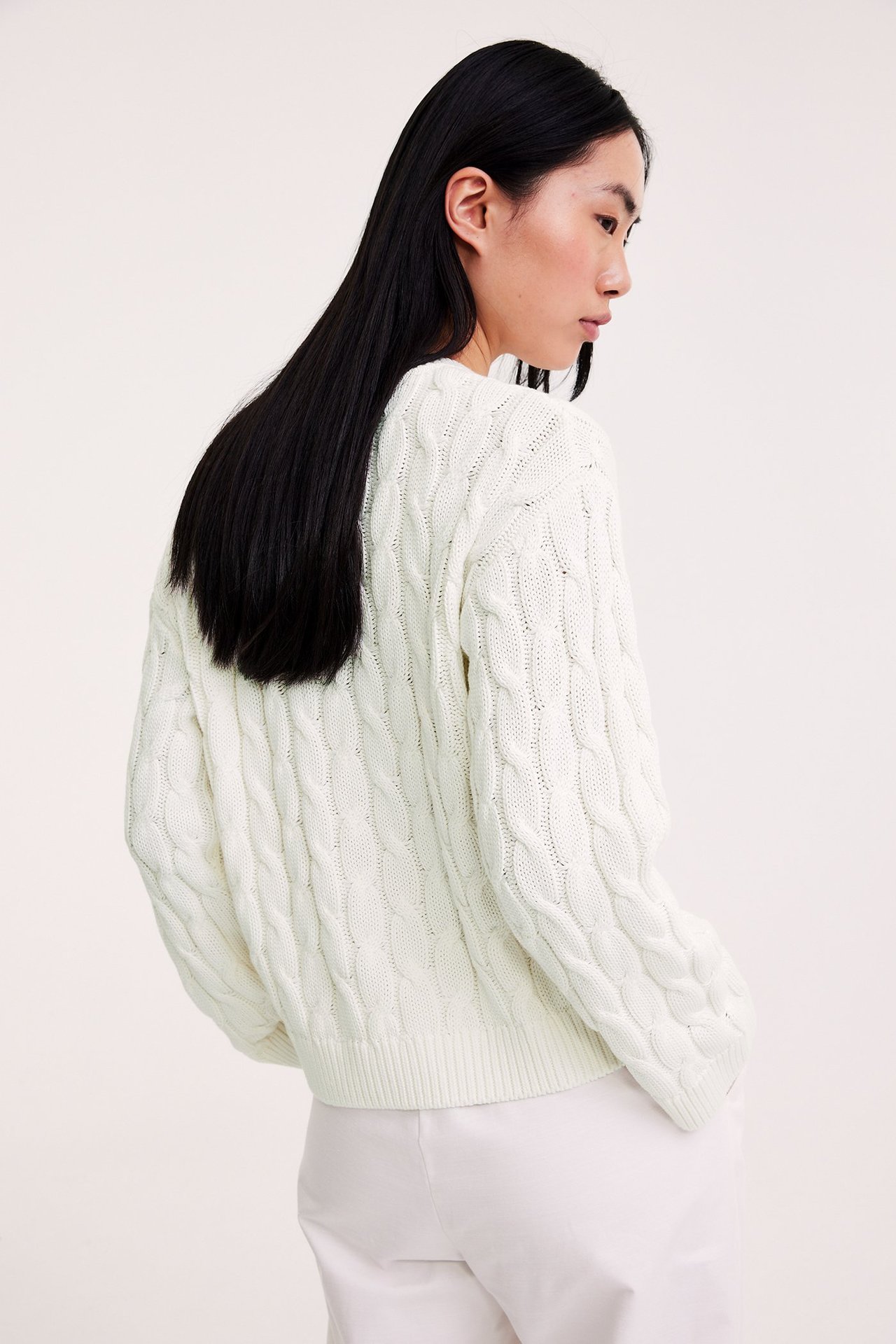 Bak-side utsikt av en kvinne i en offwhite kabelstrikket bomullsgenser og hvite bukser.