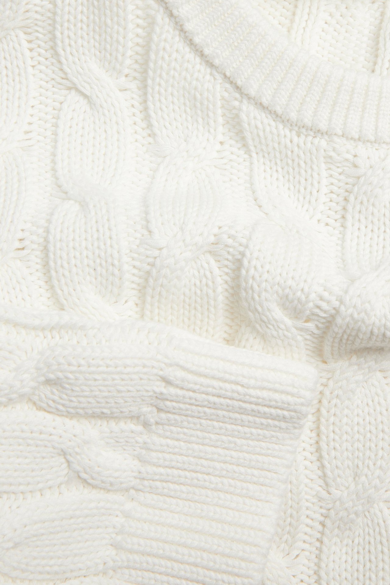 Nærbilde av en offwhite bomullsgenser med flettestrikk, ribbestrikket halskant og vrangbord.